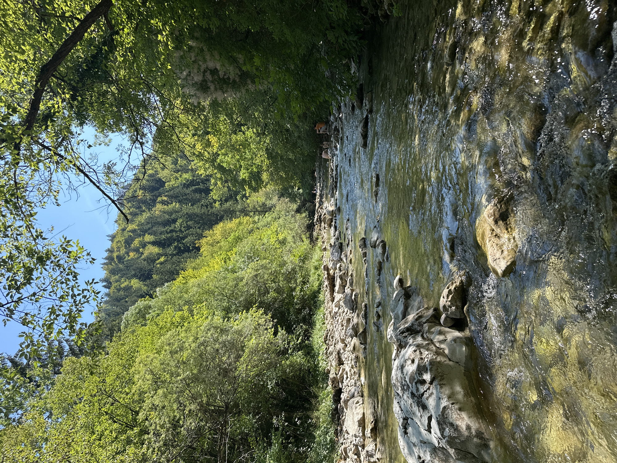 The image depicts a clear, shallow river flowing through a wooded area. The riverbed is composed of numerous stones and rocks, some of which are submerged. The water is transparent, allowing visibility of the riverbed and reflecting the surrounding greenery. Lush trees with vibrant green foliage line both sides of the river, forming a dense canopy that partially shades the water. In the background, a hillside covered with trees rises steeply. The sky is visible in the upper portion of the image, appearing clear and blue.
Further up the river, in the distance, there are three individuals. One person, wearing an orange shirt, is seated on a rock on the riverbank. Two other individuals, appearing to be unclothed, are in the river. One is standing in the water, while the other is seated on a rock. The scene suggests a natural, recreational setting, possibly a popular spot for swimming or wading. The location is identified as Iška, Slovenia.