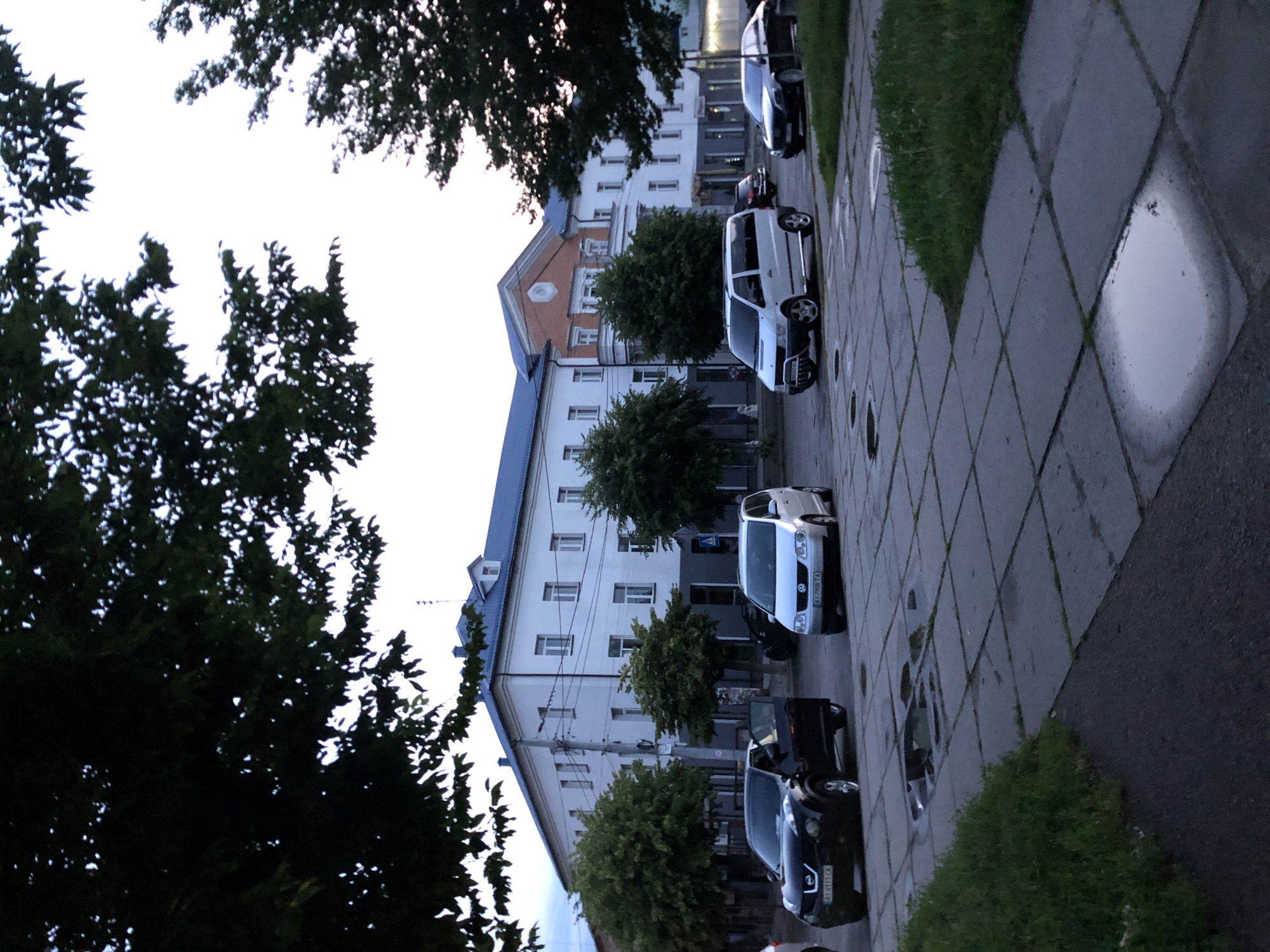 The image shows a street scene with several cars parked and driving. A white building with a blue roof dominates the background, with trees lining the street. The foreground consists of a sidewalk made of square concrete slabs, some of which have puddles of water. A black Nissan SUV with its driver's door open is parked on the left. To its right, a silver Volkswagen minivan is also parked. Further to the right, a black sedan is seen on the road. The sky is overcast, suggesting it might have recently rained. The scene appears to be in an urban environment during daytime.