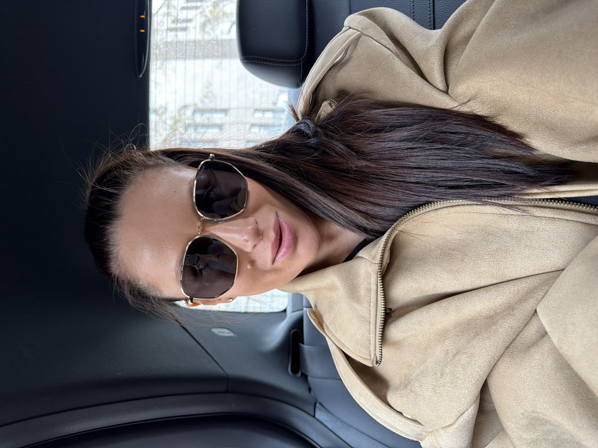 The image displays a woman seated in the back seat of a car, rotated 90 degrees clockwise from a standard portrait orientation. She is looking directly towards the camera with a subtle, relaxed smile, and her lips are adorned with a light pink gloss.
The woman has long, dark brown hair, which is pulled back and secured, with a tie visible at the back of her head. She is wearing large, gold-framed aviator-style sunglasses, with the brand "Polaroid" faintly visible on the right lens (from the viewer's perspective). Her attire consists of a light brown or tan jacket, made of a textured fabric resembling suede, with a prominent metal zipper running down the front.
The car's interior is dark, featuring black seats and a headrest. Through the side window, a blurred urban landscape with buildings is visible, suggesting the car is in a city. Small water droplets can be seen on the window pane, indicating recent rain or condensation. The scene is lit by natural daylight. The context places this scene in Moscow, Russia.