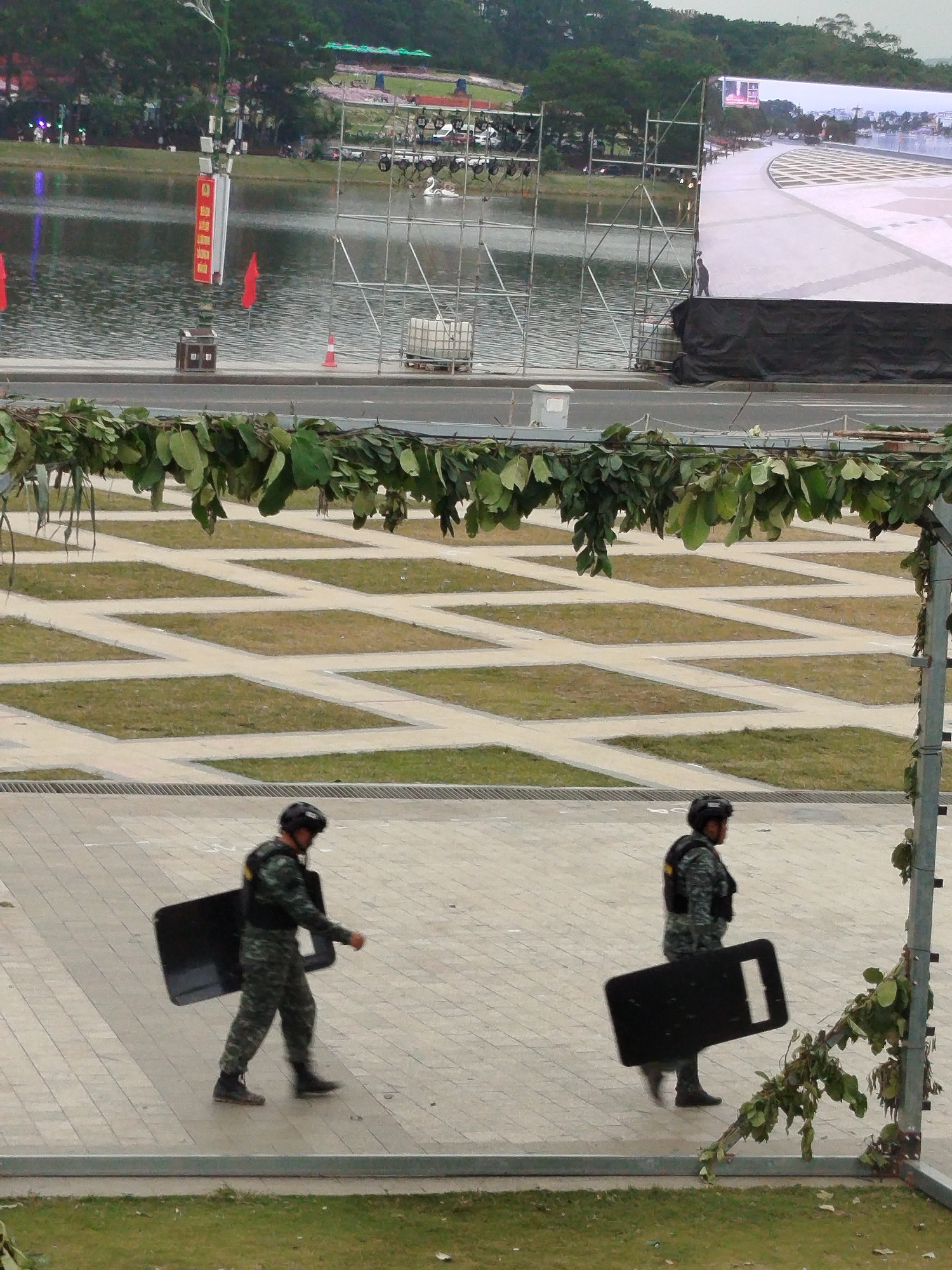 The image captures an outdoor scene near a body of water, likely a lake, in Da Lat, Vietnam. In the foreground, two individuals in camouflage uniforms and protective helmets are walking on a paved area. They are each carrying large, black riot shields. The individuals are positioned at different distances from the camera, with the one on the left closer and the one on the right further back.
Beyond the paved area, there is a section of patterned green grass interspersed with light-colored pathways. A road runs parallel to this grassy area, and behind the road is the body of water. On the water, there are several structures, including scaffolding, a large screen, and what appears to be a white swan decoration. Red flags and a tall, red and white sign are also visible near the water's edge. In the background, a forested hillside rises, with some buildings or structures visible on its lower slopes. The overall impression is of a public space, possibly set up for an event, with security personnel present.