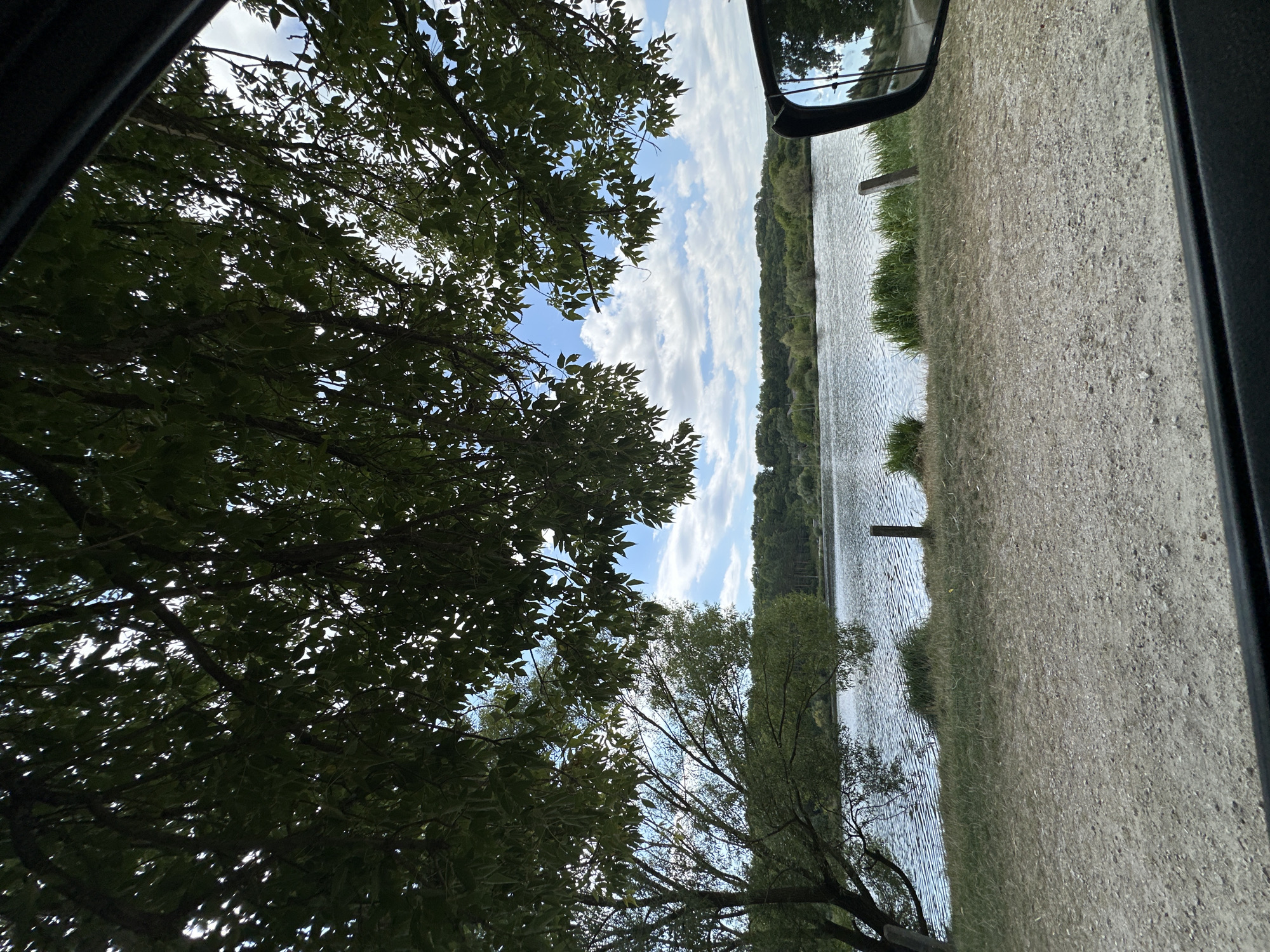The image shows a body of water, likely a lake or river, viewed from a grassy bank, with trees in the foreground and middle ground. The sky is partly cloudy and blue. In the lower right corner, a car's side mirror is visible, reflecting trees and sky. The scene appears to be a natural outdoor setting, possibly a park or recreational area within or near Makiv, Ukraine. No people are visible.