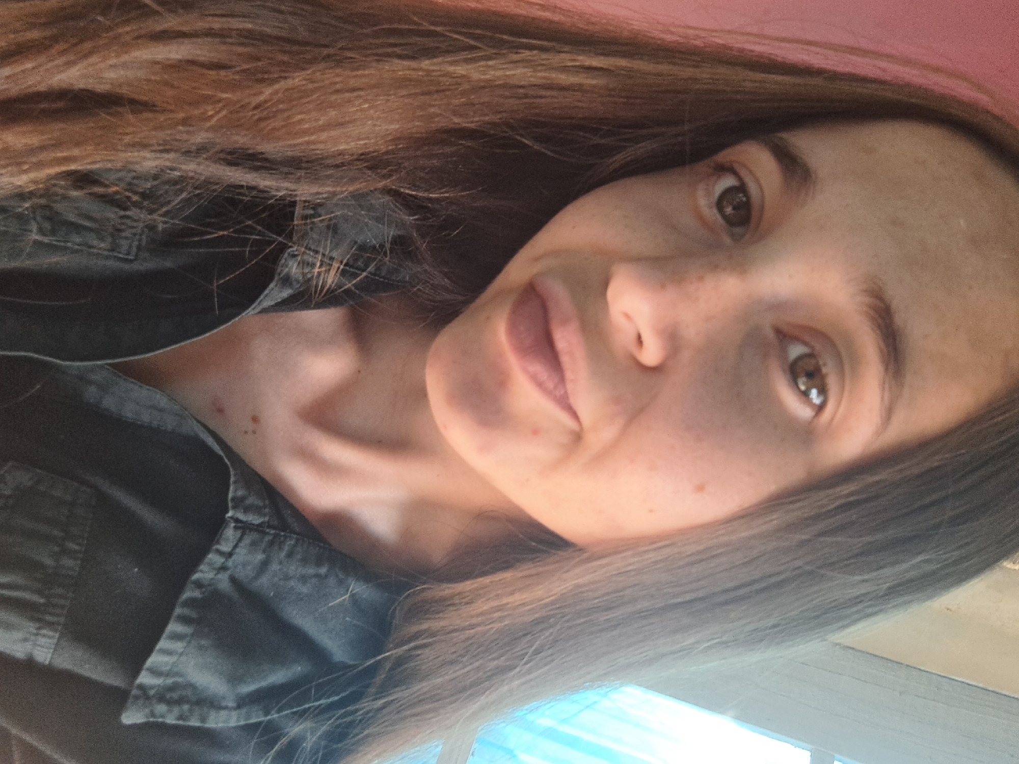 The image is a close-up, selfie-style portrait of a young woman with long, brown hair and freckles. She is wearing a dark collared shirt. Her eyes are looking towards the camera, and her lips are slightly parted. The background is softly blurred, with a pinkish hue on the left and a bright, possibly window-like, light source on the right. There are no visible landmarks or details that would suggest a specific location within the city of Vypasne, Ukraine. The scene is indoors and appears to be casually lit.