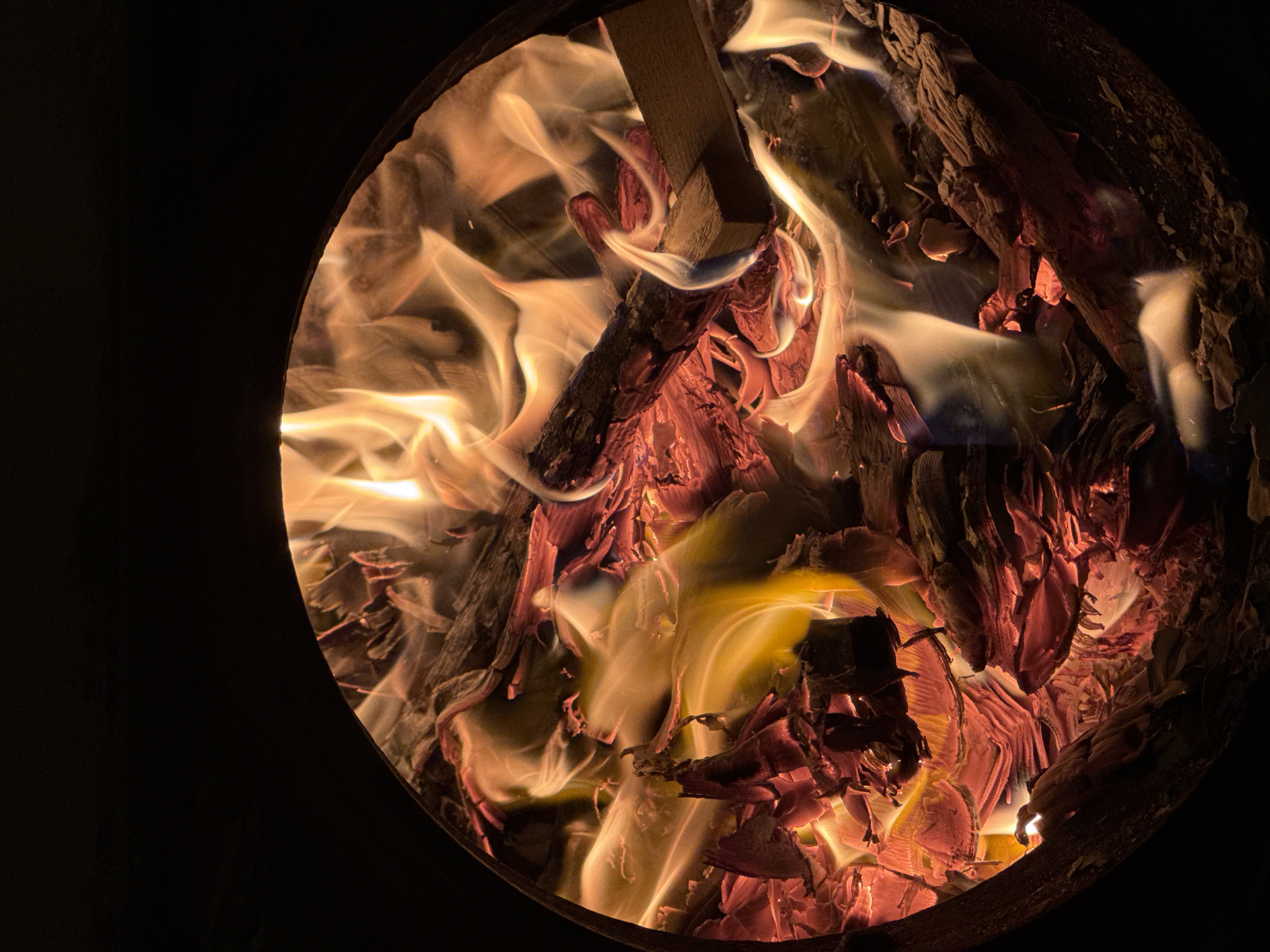 The image shows a close-up view of a bonfire contained within a dark, circular opening. Flames, in shades of yellow and orange, dance around burning wood and embers. The wood appears to be in various stages of combustion, with some pieces still intact and others reduced to glowing coals and ash. The background is entirely black, suggesting the photograph was taken in low-light conditions. There are no people or animals visible in the image. The scene is likely outdoors at night. The image does not contain any information that would allow for the identification of a specific location in Saint Petersburg, Russia.