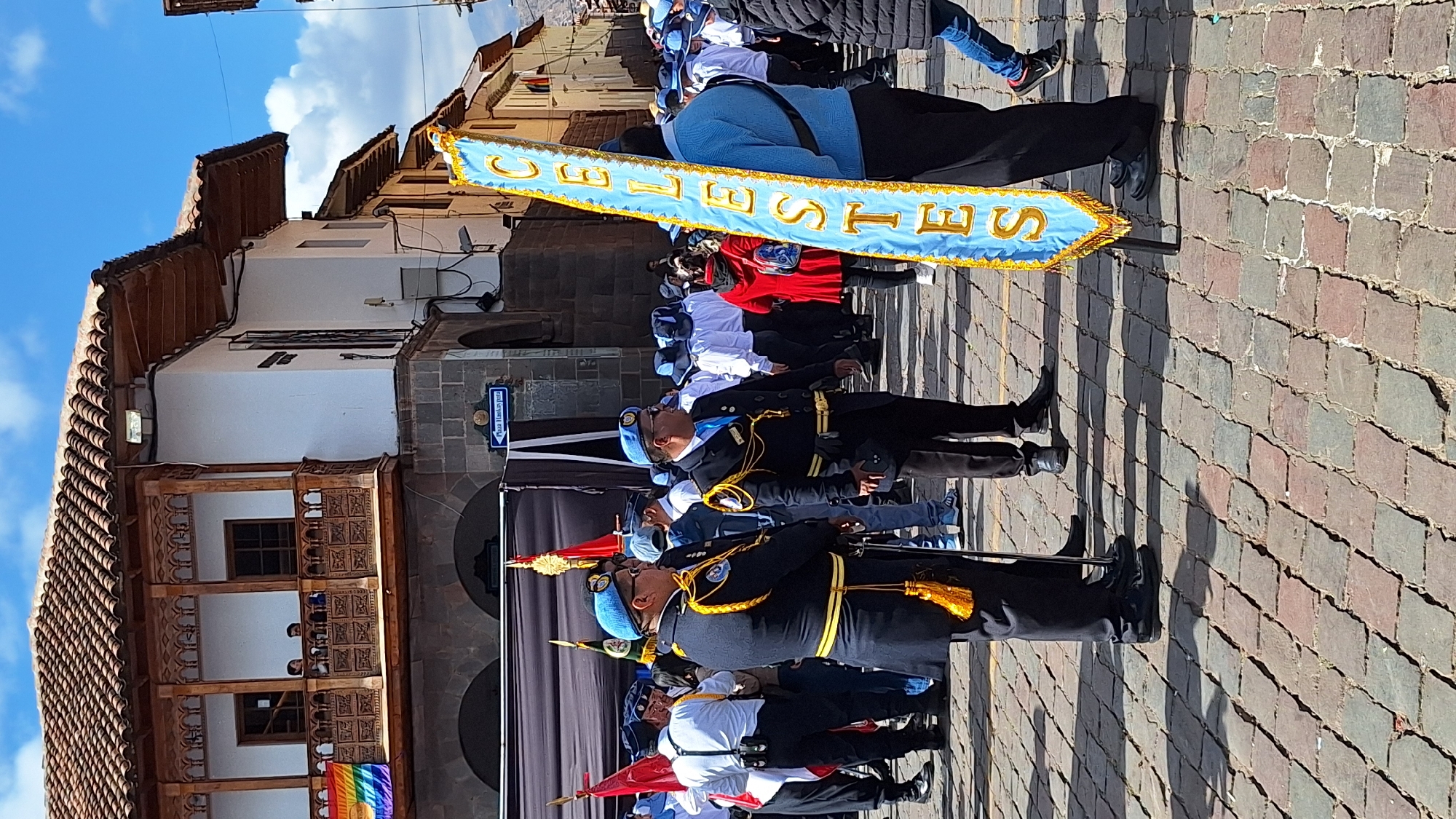 The image depicts a street scene in the city of Cuzco, Peru, likely in the Plaza de Armas, given the colonial architecture. The foreground is dominated by cobblestone paving, casting long shadows from the buildings and people present. A group of individuals, most of whom are wearing uniforms or formal attire, are gathered. Two men in the center are dressed in dark blue uniforms with light blue berets and gold trim, suggesting a ceremonial or official capacity. One of them holds a sword. To the right, a banner with the word "CELESTES" written in gold lettering on a light blue background is prominently displayed, carried by a person whose lower body is visible.
In the background, a multi-story building with a red-tiled roof and a wooden balcony adorned with intricate carvings is visible. A rainbow flag is hung on this balcony, indicating a possible Pride event or celebration. Other people are visible in the background, some wearing white shirts and hats, suggesting participation in a parade or event. A street sign with text, including "Plaza Huaskaypata," is mounted on a building. The overall atmosphere appears to be that of a public gathering, possibly a parade or a cultural event, under a bright, clear sky.