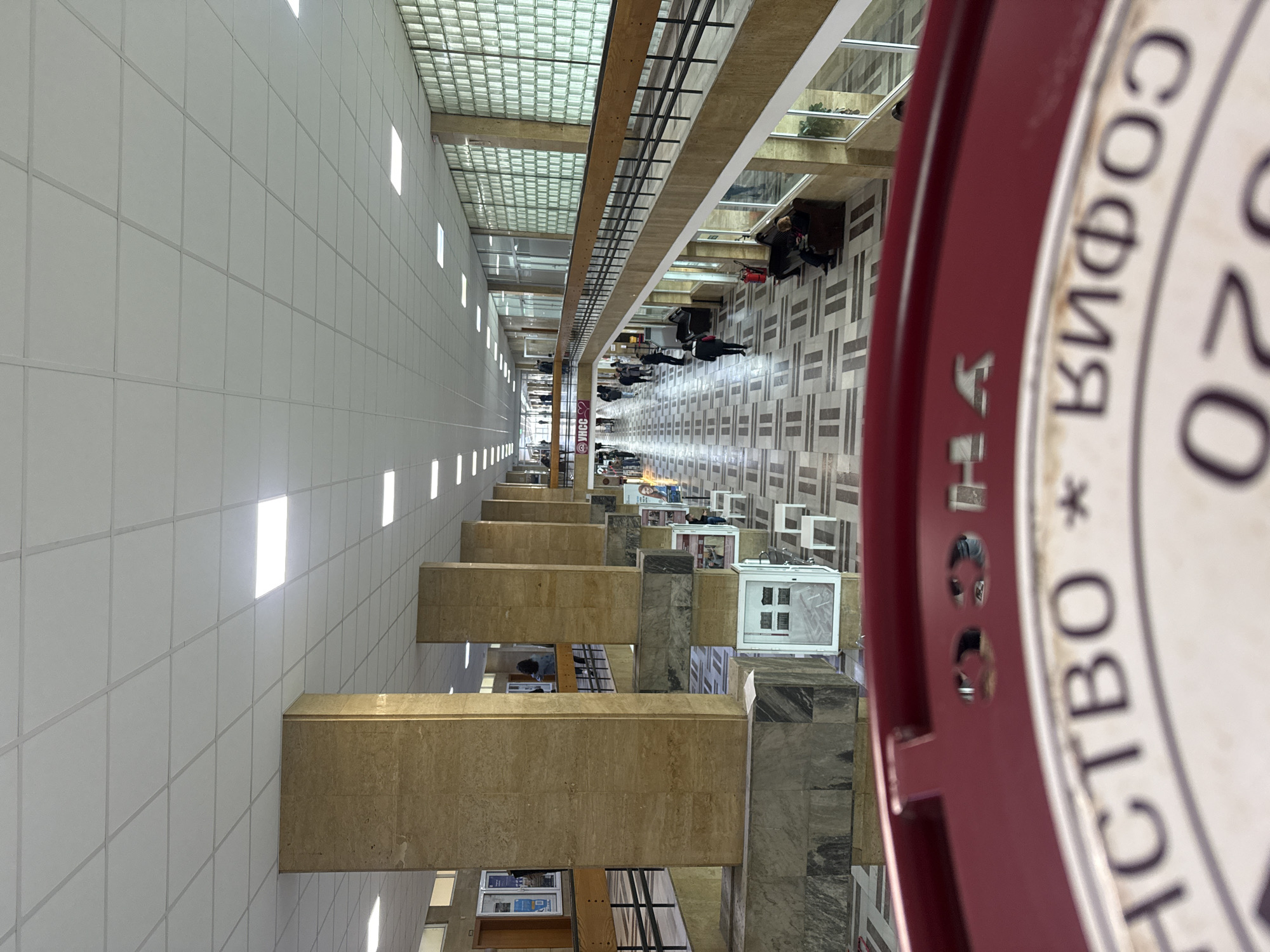 This is an interior shot of a building, likely a university or governmental building in Sofia, Bulgaria. The perspective is long and deep, suggesting a wide hallway or atrium. The scene includes architectural details like large columns that support the ceiling, and what appear to be display cases or small shops lining the hallway. People are seen walking or sitting along the hallway, and the floor has a pattern of gray and white tiles. The light fixtures on the left wall are spaced evenly. There is a sign that is partially visible and features what seems to be Cyrillic script, indicative of a location in Bulgaria, specifically Sofia, based on the visible text. The overall environment appears to be clean and well-maintained.