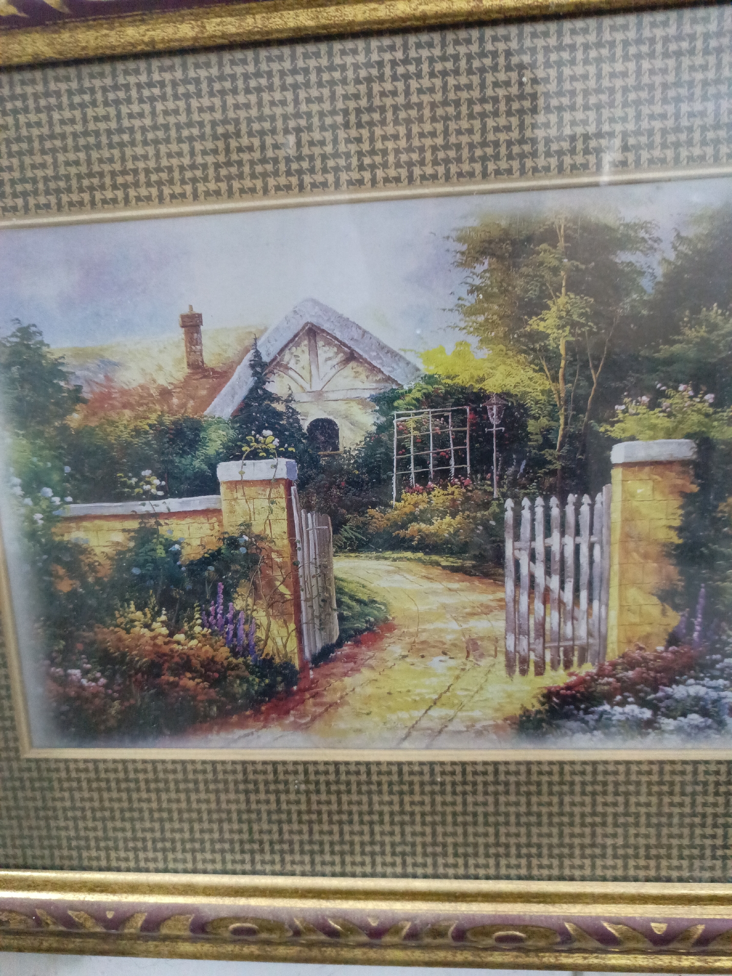 The image depicts a framed painting. The painting shows a cottage scene with an open white picket fence leading up a path to a cottage with a thatched roof. The path is surrounded by various flowers and greenery. A trellis is visible in the background. The painting is enclosed in a gold and patterned frame.
The location is not Juan Griego, Venezuela. The image has no relation to that city.