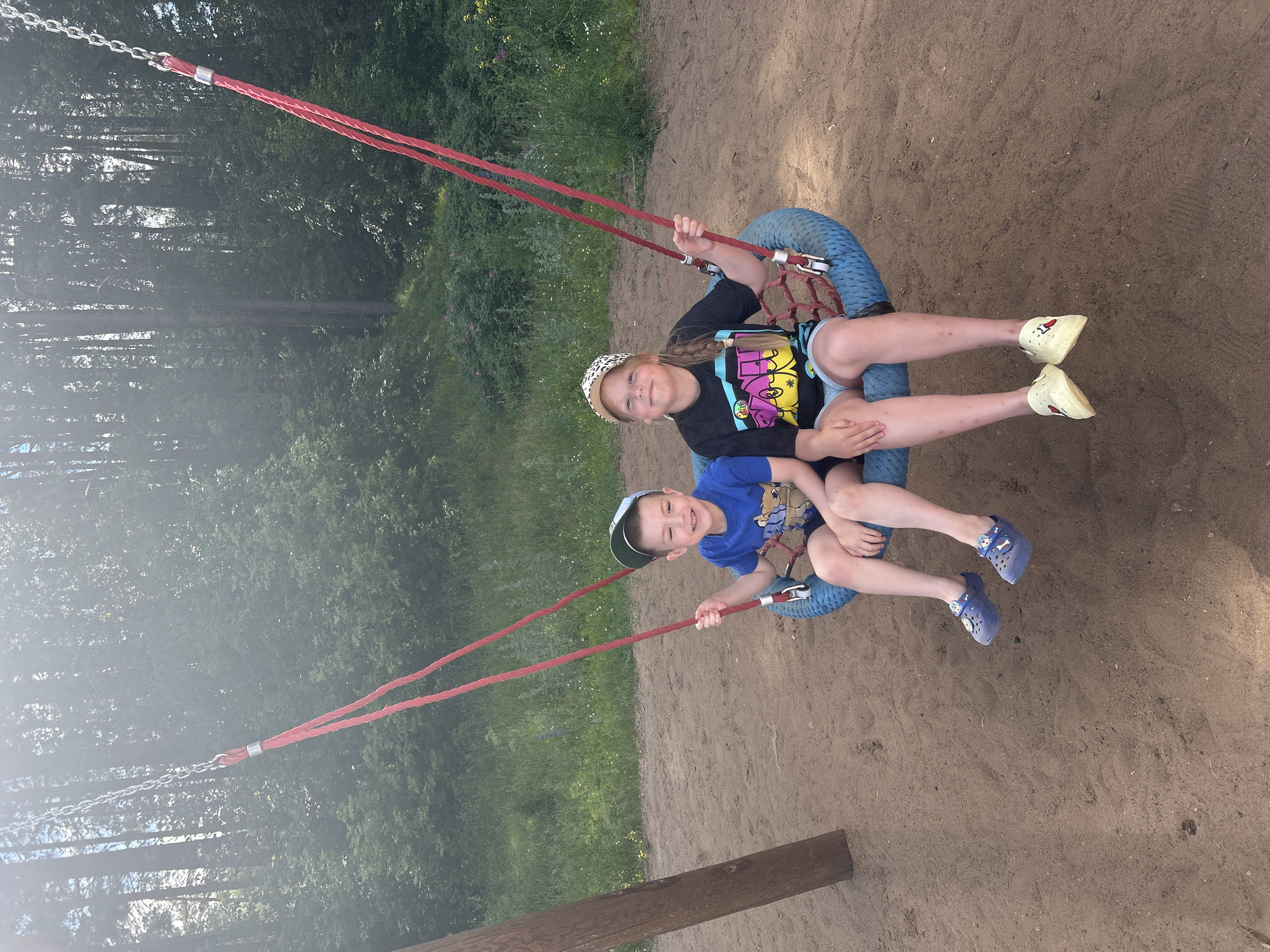 Two children, a boy and a girl, are seated on a tire swing in a sandy playground. The boy, on the left, wears a blue t-shirt with a cartoon character graphic, dark shorts, a green and blue baseball cap, and blue Crocs. He is smiling and looking to his right. The girl, on the right, has blonde hair in a braid and wears a black t-shirt with a colorful graphic, light blue shorts, a patterned baseball cap, and yellow slip-on shoes. She is also smiling and looking at the camera. The tire swing is suspended by red ropes and metal chains from a wooden post, partially visible on the left. The background consists of a dense forest with tall trees and green foliage, with a grassy area in front of the trees. The ground of the playground is composed of sand. The image appears to be taken on a sunny day, with some lens flare visible in the upper part of the frame. The scene is located in a park or outdoor recreational area within the city of Chaikovsky, Russia, likely a public playground.