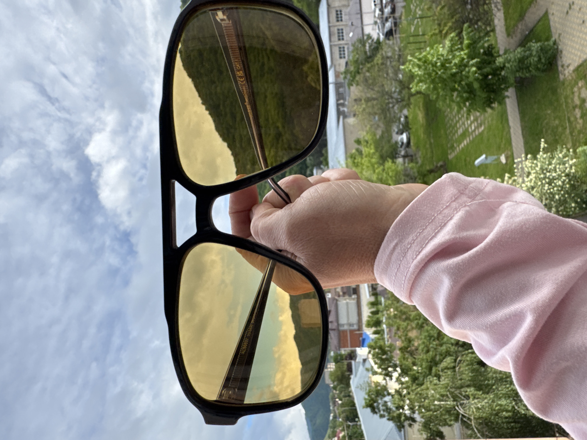 A close-up shot features a pair of oversized sunglasses with dark frames and yellow-tinted lenses, held by a fair-skinned hand wearing a light pink long-sleeved shirt. The reflection in the glasses shows a landscape, including a bridge, hills, and buildings.
The background outside the lenses shows a cityscape with greenery, buildings, and cars, typical of an urban environment. The sky is visible in the upper left corner, suggesting an outdoor setting during daylight.
The location is in Karachayevsk, Russia.
