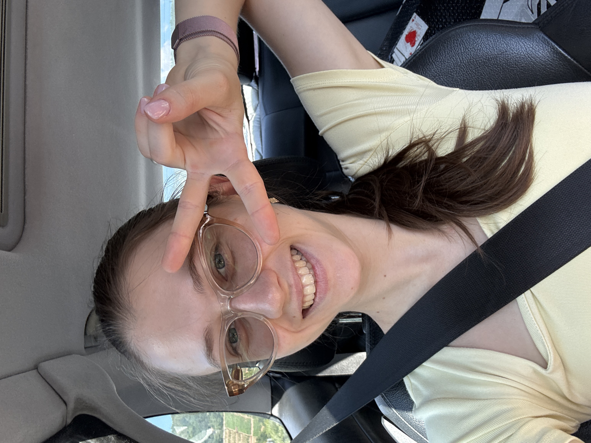 A young Caucasian woman with light skin, light brown hair pulled back, and light green eyes is in the driver's seat of a car, looking at the camera and making a peace sign with her right hand. She is wearing clear-framed glasses and a pale yellow top with a low neckline. A black seatbelt is diagonally across her chest. The background shows the interior of the car and a blurry view of trees and fields through the window. The location is not Wörth am Main, Germany.