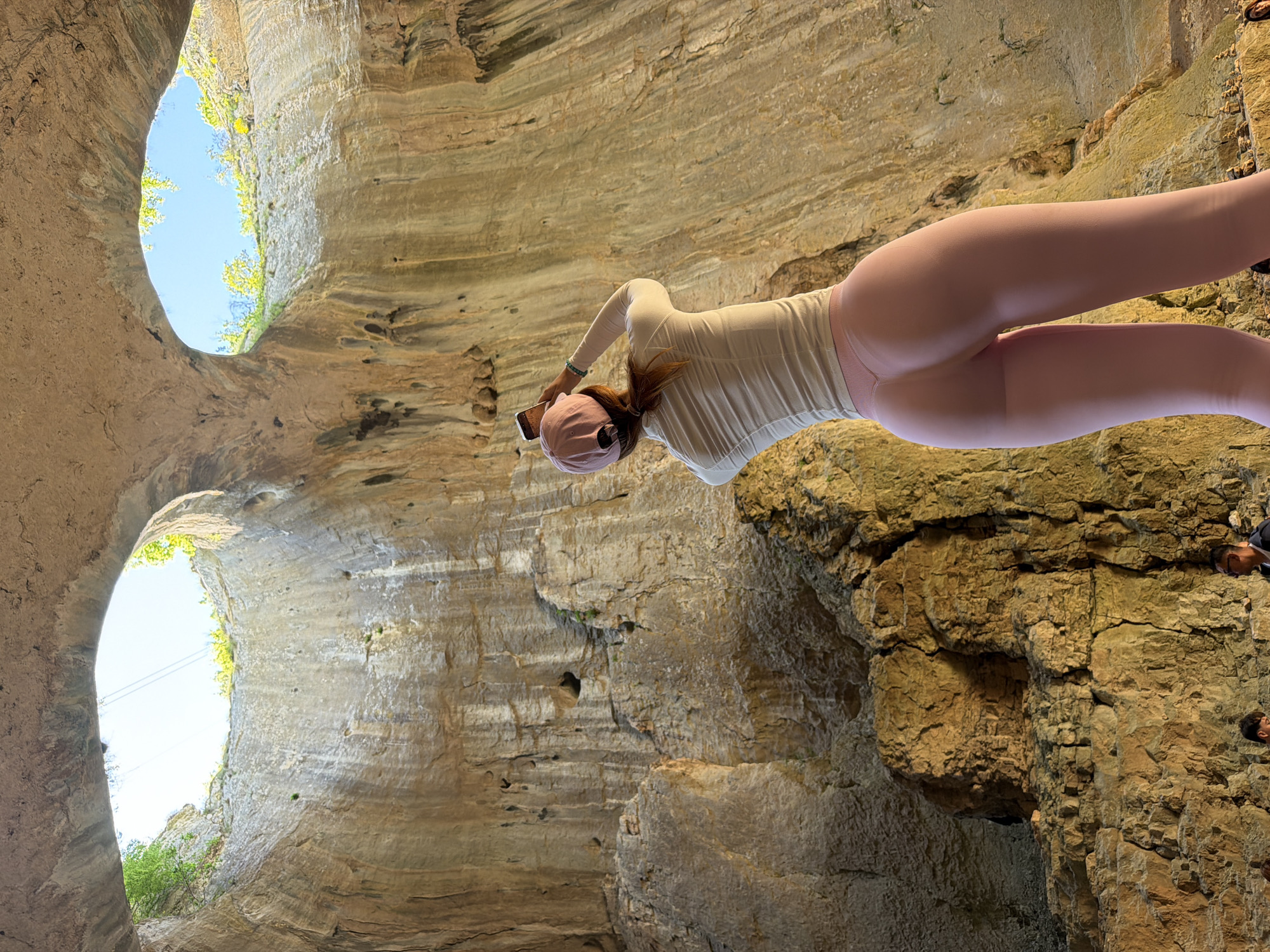 The image depicts a person standing within a cave structure, likely a part of the Devetashka cave system, near Karlukovo, Bulgaria. The person is facing towards the cave wall, holding a mobile phone in their left hand. The person is wearing a long-sleeved light-colored top, pink leggings, and a pink baseball cap. The cave has layered, sedimentary rock formations, with two large openings at the top allowing light to enter. Vegetation is visible through the openings. The background consists of the rocky walls and the bright sky visible through the cave openings. The person appears to be taking a photo or recording a video of the cave interior.
