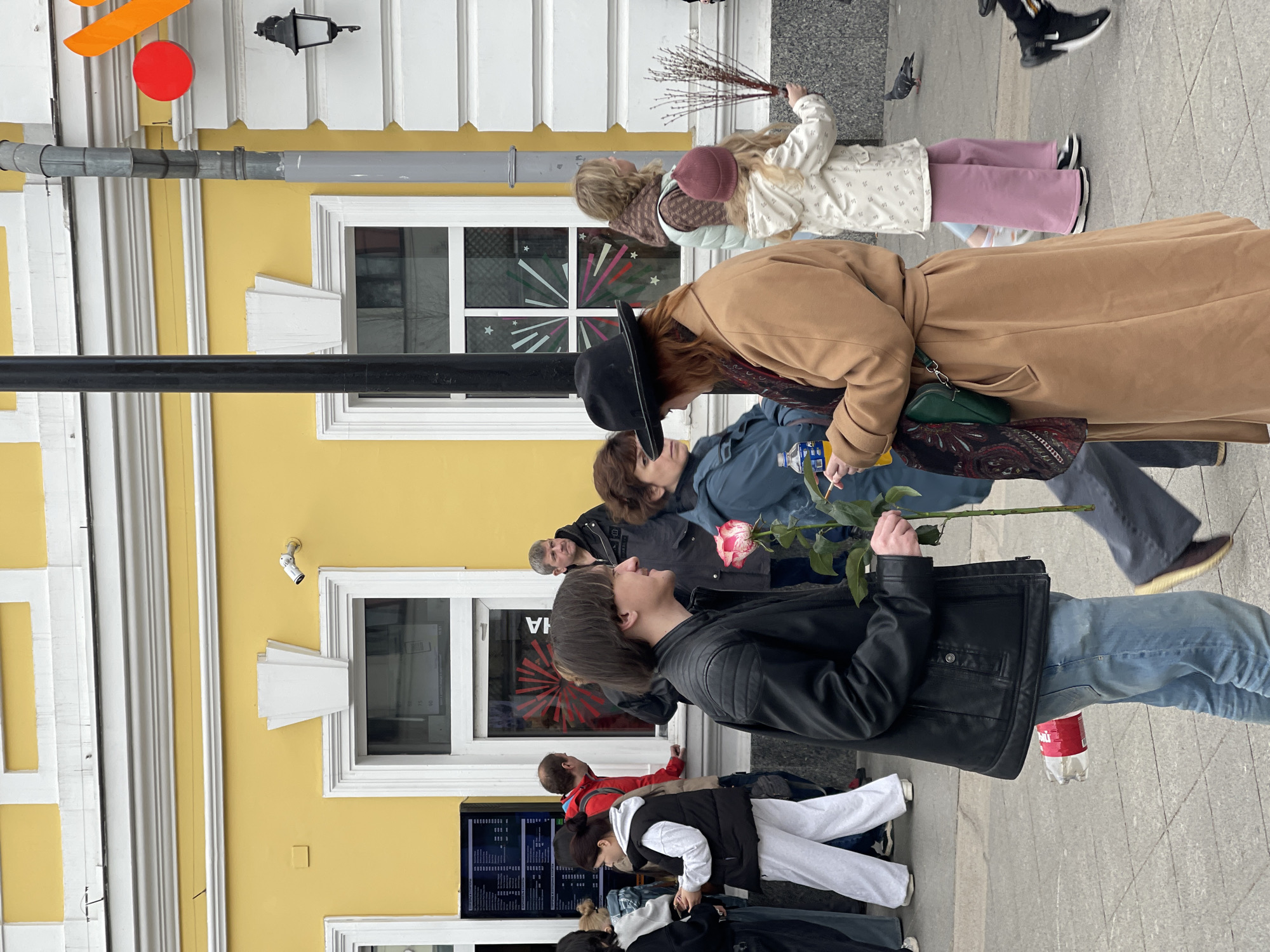 A group of people are standing on a sidewalk near a yellow building in what appears to be a city environment. The people seem to be waiting or engaged in some activity, with some holding items like flowers and branches. The building has windows, white frames, and a menu board. The location may be Moscow, Russia, based on architectural style and the presence of Russian text.