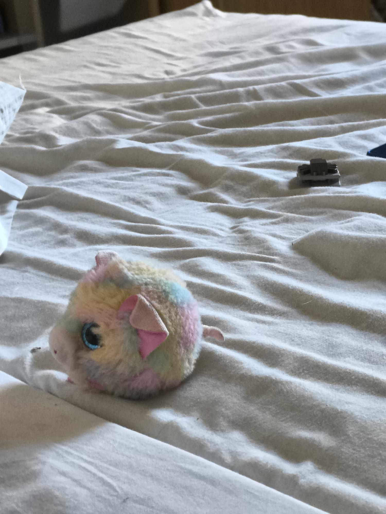 The image shows a bed covered in a white, wrinkled sheet. A plush, multi-colored pig toy with blue eyes and pink ears is positioned on the left side of the bed. Towards the upper right of the image, a small gray object, possibly a toy car or vehicle component, is visible. The scene is indoors. The location in Pélussin, France is impossible to determine from the information provided.
