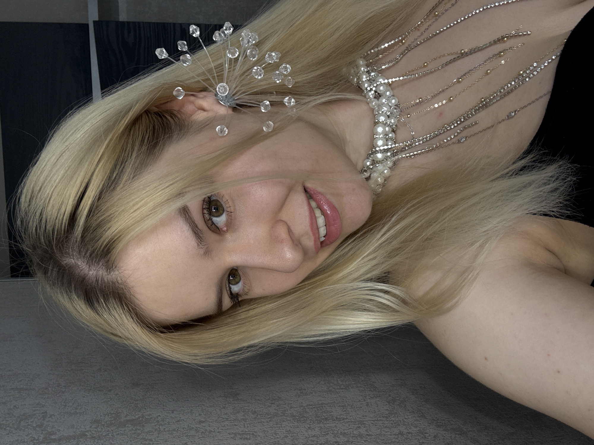 A medium shot shows a young woman with long, blonde hair and light brown eyes. She is wearing a black top, a pearl necklace, and a large, crystal-embellished earring on her right ear. Her head is tilted to her left, and she is looking at the camera with a slight smile. The background is a textured gray wall on the left and a dark, reflective surface on the right. The lighting is soft, highlighting her face and jewelry.