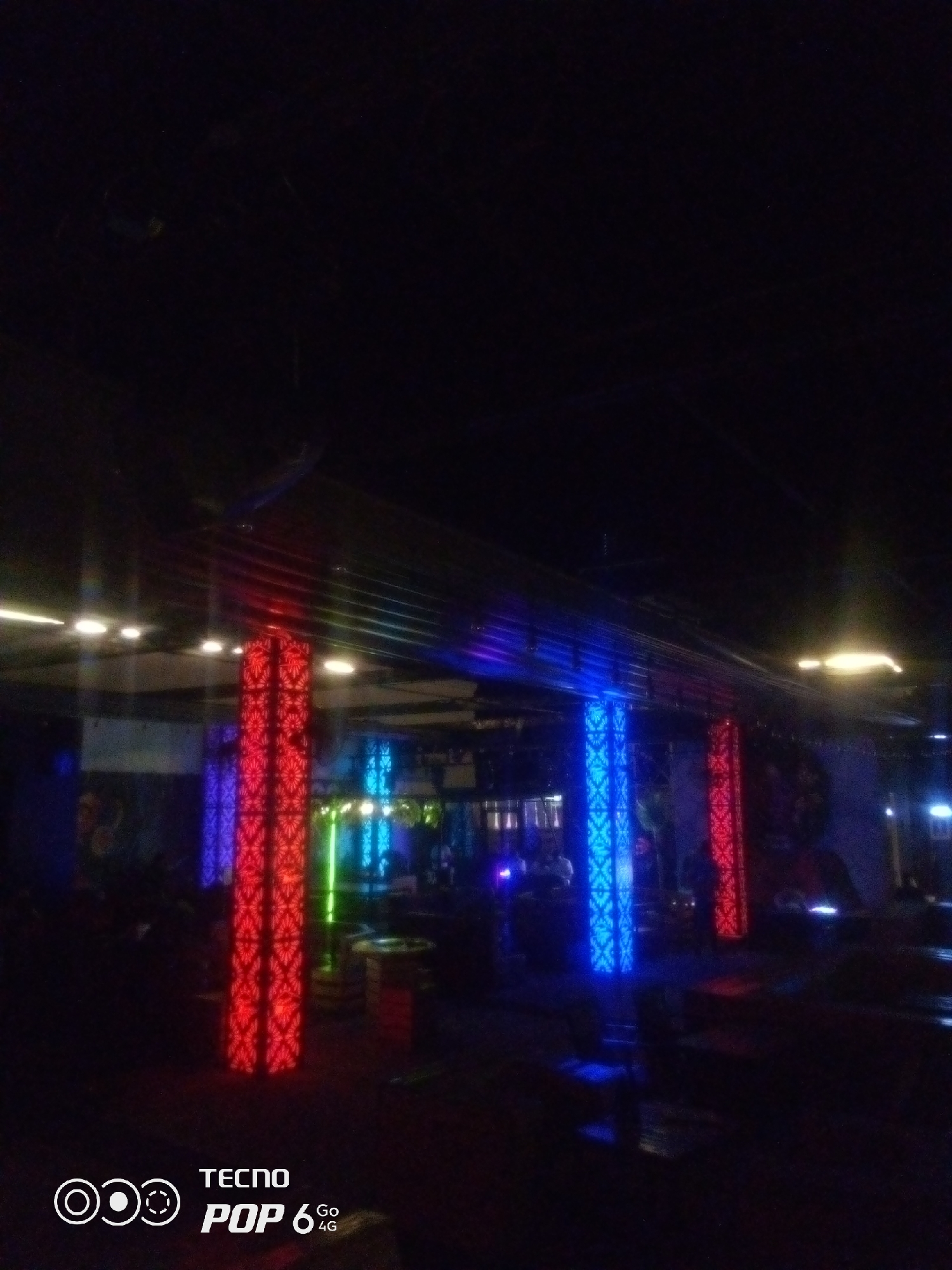 The image shows the interior of a dimly lit establishment, likely a bar or club, in Howayi, Nigeria. The primary illumination comes from decorative LED lights on pillars. Two prominent pillars are visible, one with intricate red lighting and another with blue lighting. Several other pillars with similar lighting patterns are visible in the background, along with some ceiling lights. The environment appears to be a public space with furniture and possibly other patrons, though they are mostly indistinct in the low light. The bottom left corner of the image features a watermark indicating "TECNO POP 6 Go 4G".