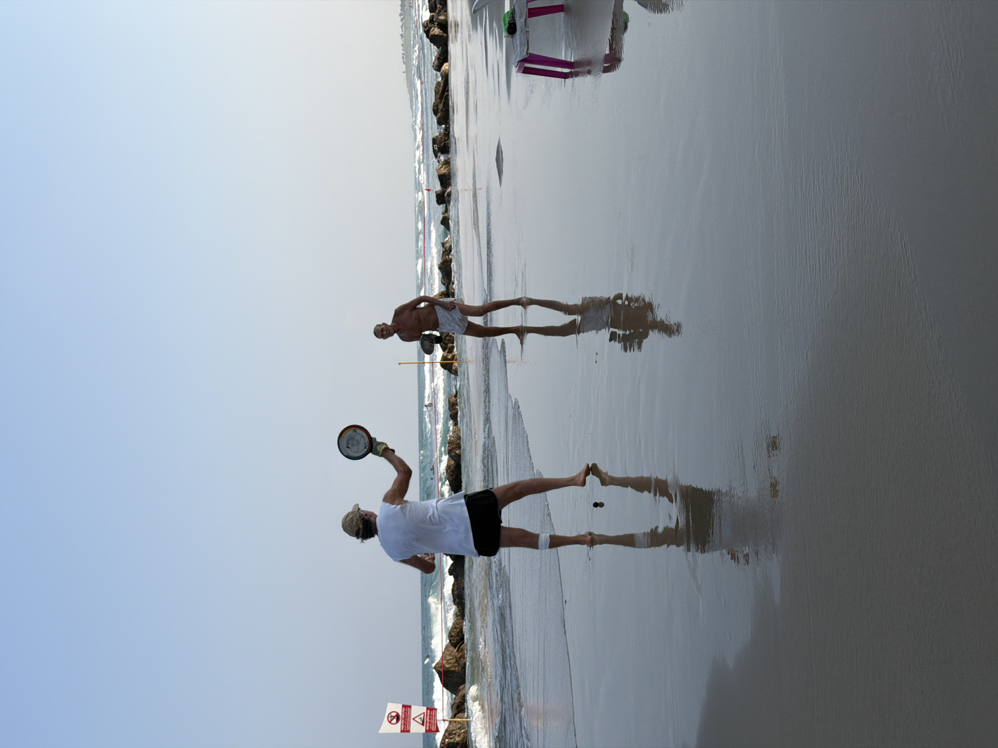 Two individuals are playing paddleball on a wet sandy beach in Bat Yam, Israel. The person in the foreground, viewed from the back, is wearing a white t-shirt, black shorts, and a beige cap. They are holding a paddle in their right hand, raised as if to strike a ball. A white bandage is visible on their left knee. Their reflection is clearly visible on the wet sand. To the right and further back, another individual, a man, is facing the camera and also holding a paddle. He is shirtless and wearing light-colored shorts. His reflection is also visible. In the background, waves are breaking on the shore, and a rocky breakwater extends into the sea. Two red and white warning signs are visible on the left side of the image, near the water's edge. A small pink table with a green towel is situated on the right side of the frame. The sky is clear and blue, with a hazy horizon.