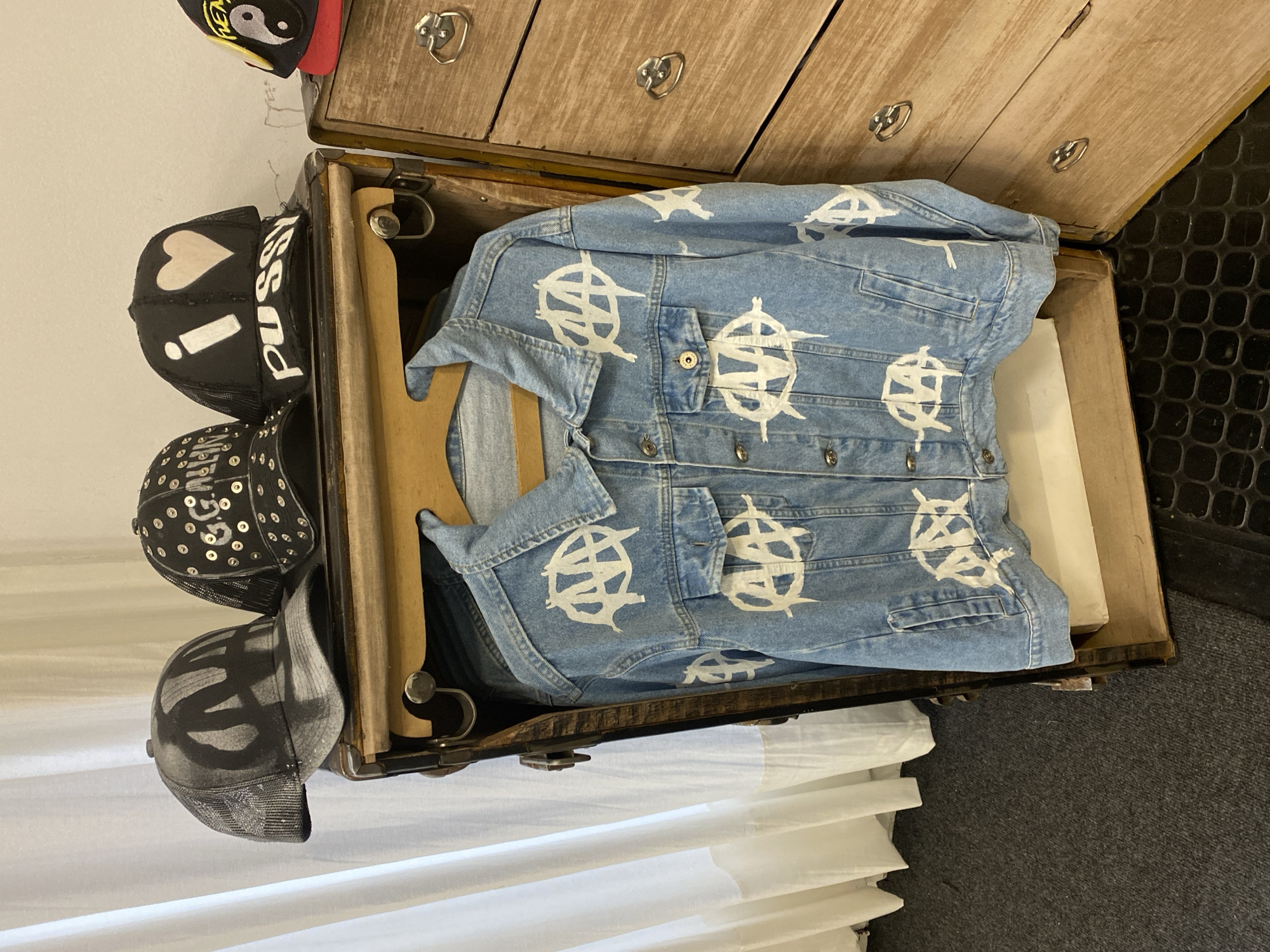 This image captures an indoor scene featuring clothing and accessories, likely within a room or storage area. The central object is a light-washed denim jacket, folded over a wooden hanger and placed inside an open, vintage wooden trunk or large suitcase. The jacket is distinctive, covered with multiple white, stylized "A" symbols, resembling either the Vlone logo or an anarchist symbol.
Above the trunk, a rustic wooden dresser with three drawers and antique-style metal ring pulls is visible. To the left of the trunk, a stack of four trucker-style baseball caps hangs against a white wall. The topmost cap is black with a yellow Yin-Yang symbol. Beneath it, a black cap features a white heart, a white exclamation mark, and the word "Pussy" written in white script along its side. The third cap is black with silver studs or sequins, and the bottom cap is black with a large, faded grey or black graphic design.
The background includes a white wall, a dark textured floor on the right, and white curtains or blinds on the left. No activity is taking place, and no people are present. The scene depicts a static arrangement of personal items. While the provided context specifies Buenos Aires, Argentina, there are no visual cues within the image itself to confirm this location.
