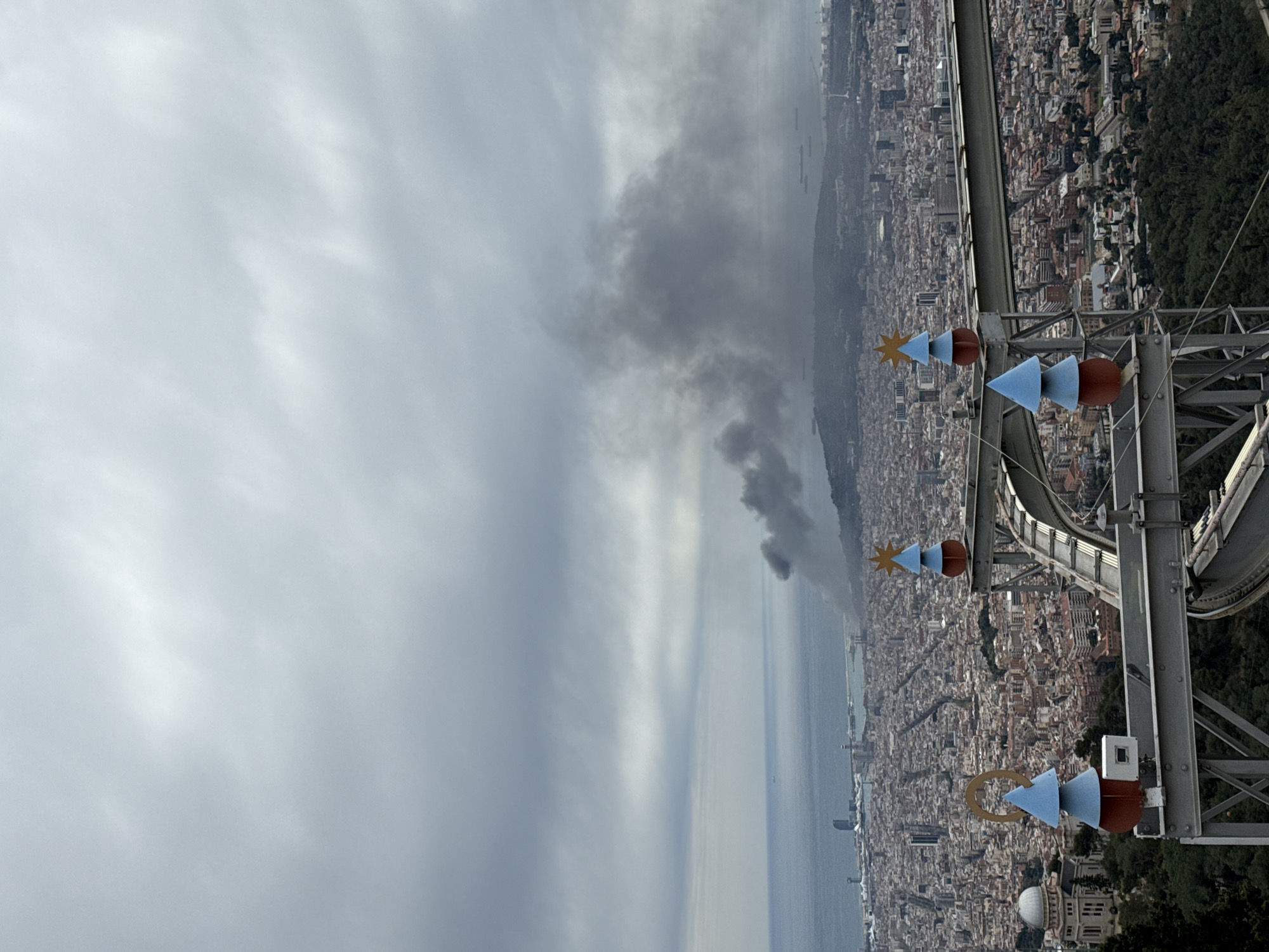 From a high vantage point on Montjuïc in Barcelona, Spain, a view overlooks the city and the Mediterranean Sea. In the foreground is the Teleferic cable car structure with colorful, stylized wind direction indicators. The city sprawls densely below, with buildings extending to the coastline. Out on the sea, a plume of dark smoke rises, possibly indicating a fire or incident. The sky is overcast with varying shades of gray clouds.