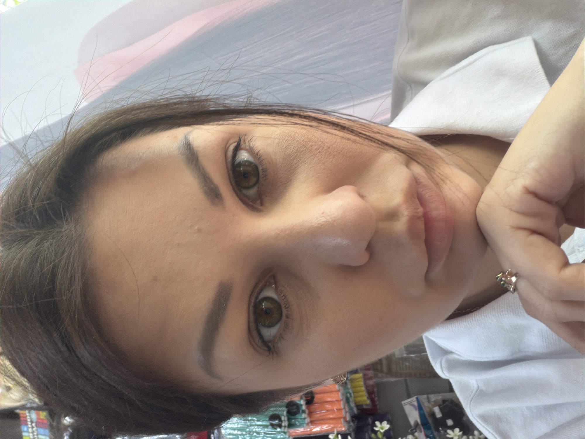 The image is a close-up selfie of a woman with brown hair and brown eyes. She is wearing a white polo shirt and a ring on her left hand, which is resting on her chin. The background is blurred and appears to be a store or market with racks of colorful items. The lighting suggests it is daytime. The location is identified as Sochi, Russia.