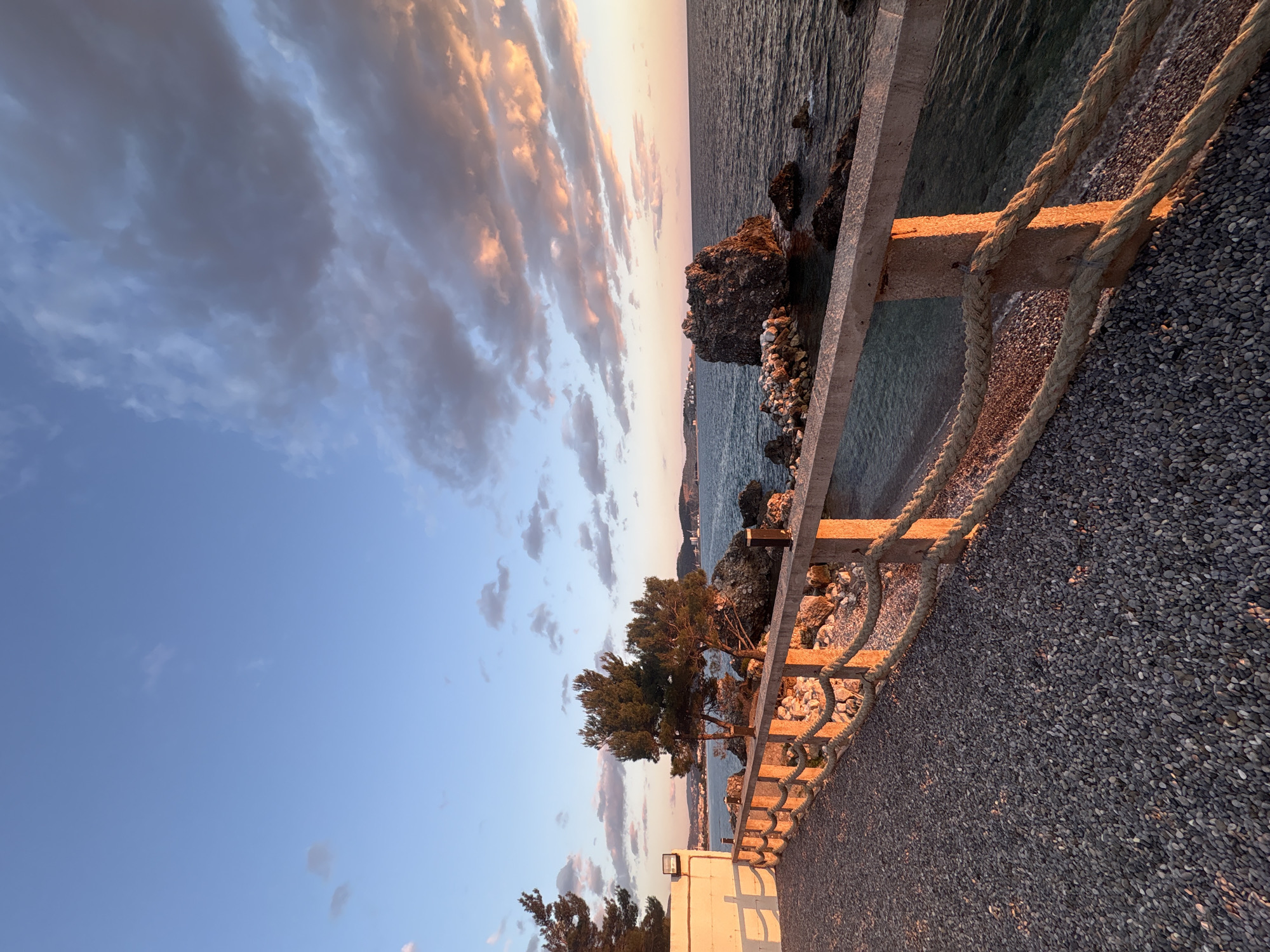 The image shows a coastal scene at sunset, possibly in the city of Bar, Montenegro. The view is from a pebble-covered promenade bordered by a concrete and rope railing. Beyond the railing are rocks and vegetation, followed by the sea and a sky filled with clouds tinged with orange and pink from the setting sun. The horizon is visible in the distance, indicating an open water view. There are trees and a small white structure near the railing. No people are visible.