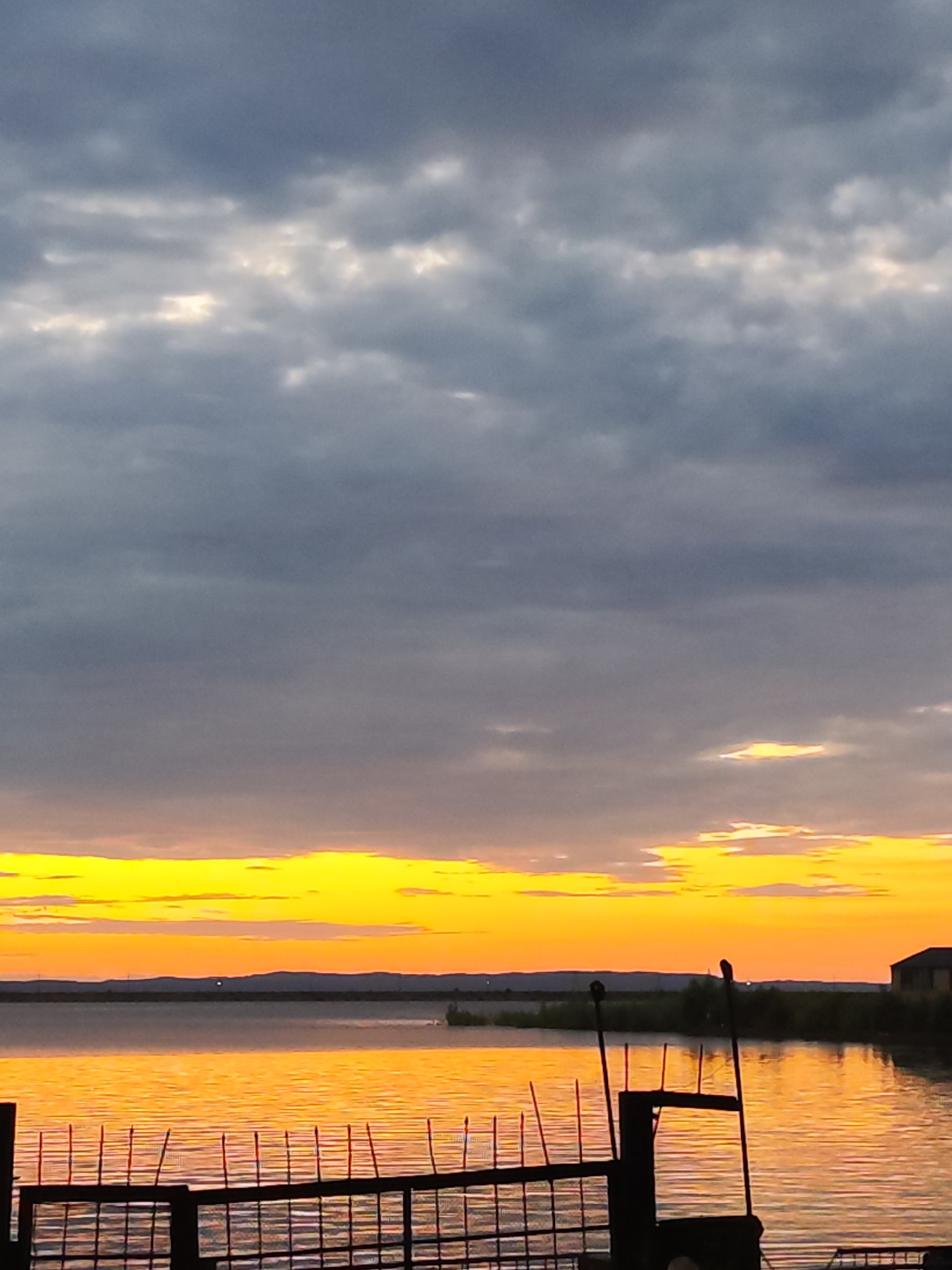 The image captures a serene sunset over a body of water, likely the Kama River, near Chaikovsky, Russia. The sky is a canvas of dramatic clouds, with dark gray formations dominating the upper portion, contrasting sharply with the vibrant bands of yellow and orange near the horizon. The setting sun casts a warm, golden glow across the rippling water, creating a reflective surface that mirrors the sky's fiery hues.
In the foreground, a dark metal fence with vertical posts is silhouetted against the bright water. Beyond the fence, the far bank of the river is lined with low-lying vegetation and trees. A single, dark structure, possibly a building, is visible on the right side of the distant shoreline. A small, dark silhouette, possibly a bird, is perched on one of the fence posts in the mid-ground. The overall atmosphere is peaceful and tranquil, characteristic of a calm evening by the water.
