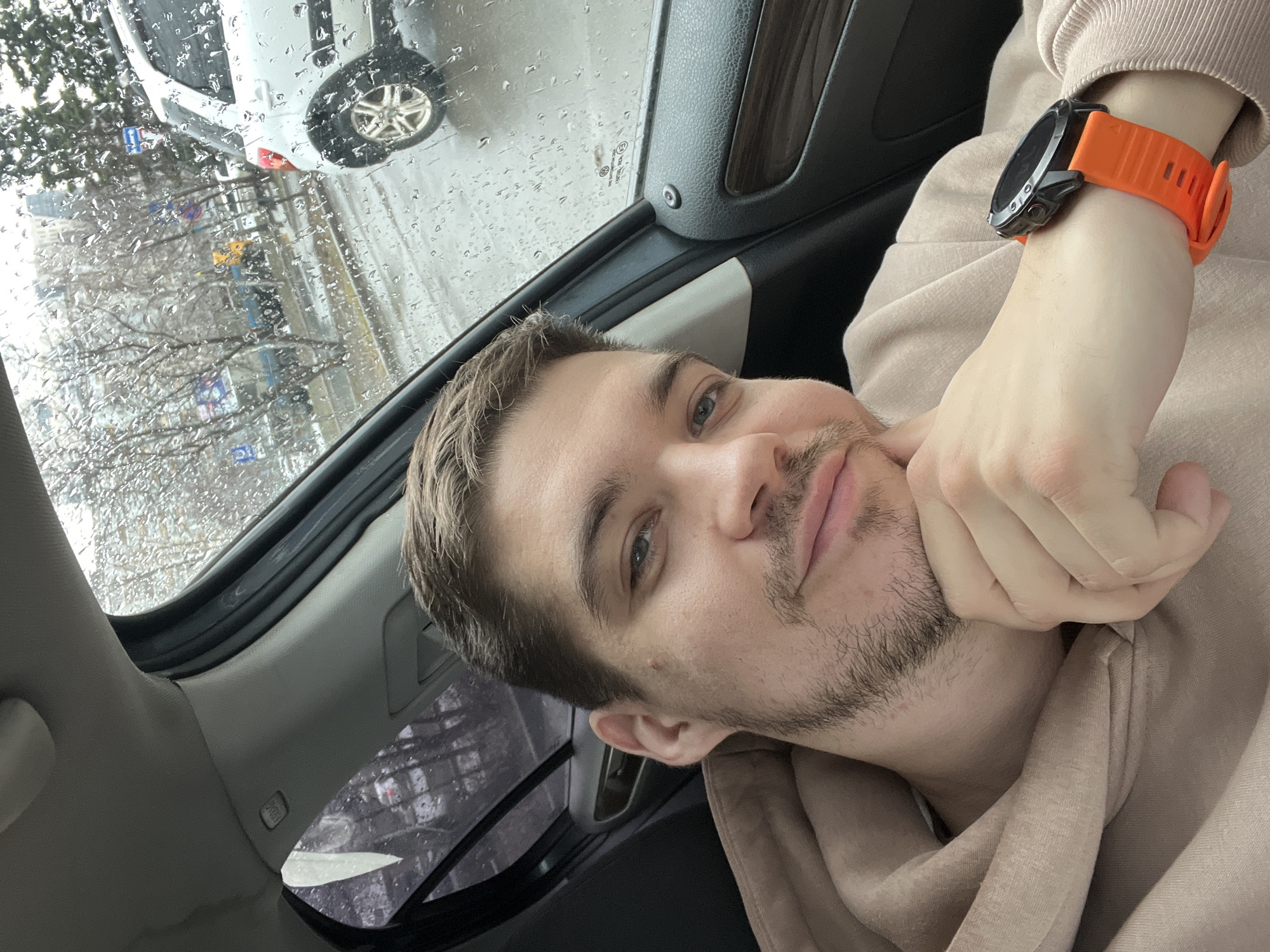 A man is inside a car, presumably in Kazan, Russia based on the Cyrillic text visible in the background outside the rain-streaked window. The man has short brown hair, light eyes, and light stubble. He is wearing an orange fitness tracker watch on his left wrist. His face is turned towards the camera, and his hand is resting on his chin. He appears to be wearing a beige or light brown hooded sweatshirt. Outside the car window, there is a building and trees, and a white car is also visible. The weather appears to be rainy.
