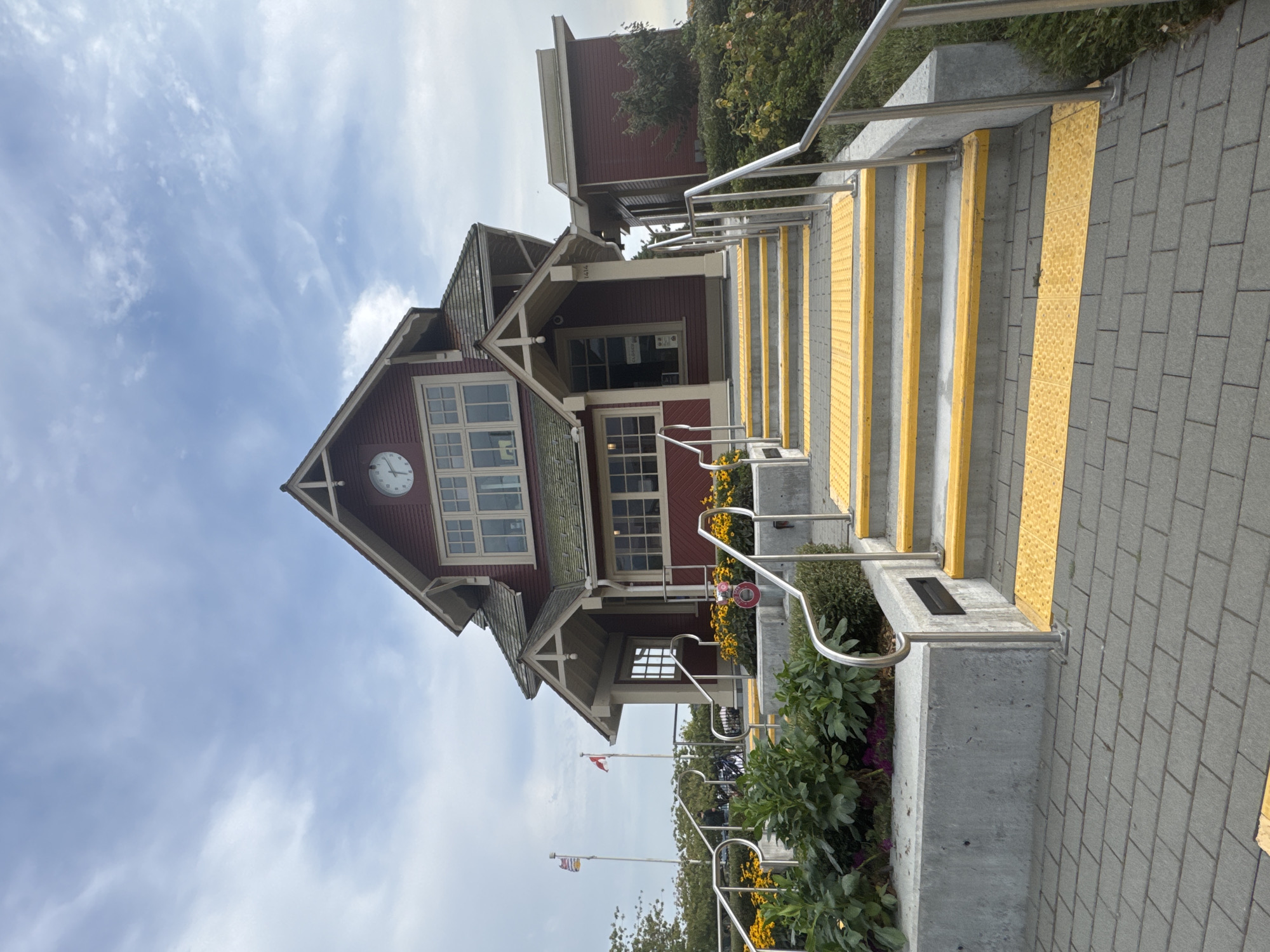 This is a ground-level, eye-level, medium shot of the exterior of the Horseshoe Bay ferry terminal building in West Vancouver, Canada. The building is painted a deep red with cream-colored trim and has a steeply pitched roof. A clock is visible on the upper portion of the building. A wide set of concrete stairs with yellow tactile warning strips and metal handrails leads up to the entrance of the building. There are yellow flowers planted on either side of the stairs. A paved walkway made of gray interlocking bricks is in the foreground. Two flags are visible in the background, one a Canadian flag and another flag of British Columbia. The sky is overcast with scattered clouds. The image is devoid of people or any discernible activity.