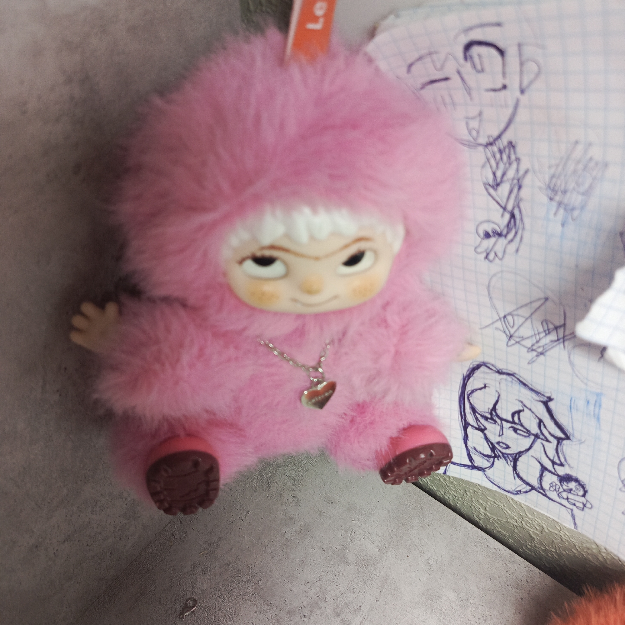 A close-up shot depicts a pink, furry doll with a human-like face, a silver necklace with a heart-shaped pendant, and brown shoes. The doll rests on a gray surface next to a piece of graph paper with various cartoon sketches in blue pen. Part of an orange object is visible in the lower right corner.