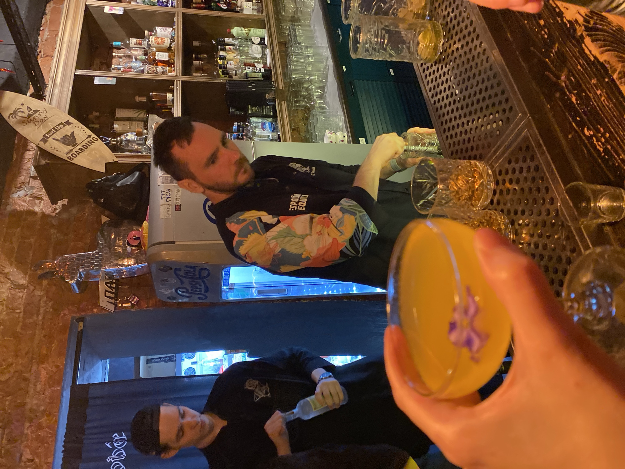 The scene is an indoor bar setting in what appears to be Izhevsk, Russia, evidenced by the Cyrillic script seen on a cooler stating "Лучшие гуси 100% НАТУРАЛ" and "Дибёр" partially obscured by a curtain. A bartender with a beard, wearing a black shirt with floral print sleeves and the partial word "ESPON" on the shirt, is pouring liquid from one glass to another. A second bartender in a black shirt with an insignia on it is pouring liquid from a bottle. A hand holds a yellow cocktail with a violet flower garnish. A bar top is visible with multiple glassware. Shelves of alcoholic beverages line the background. Wall decor includes a surfboard and a Pepsi cooler with the phrase "EPS 8 800 700 00 55".
