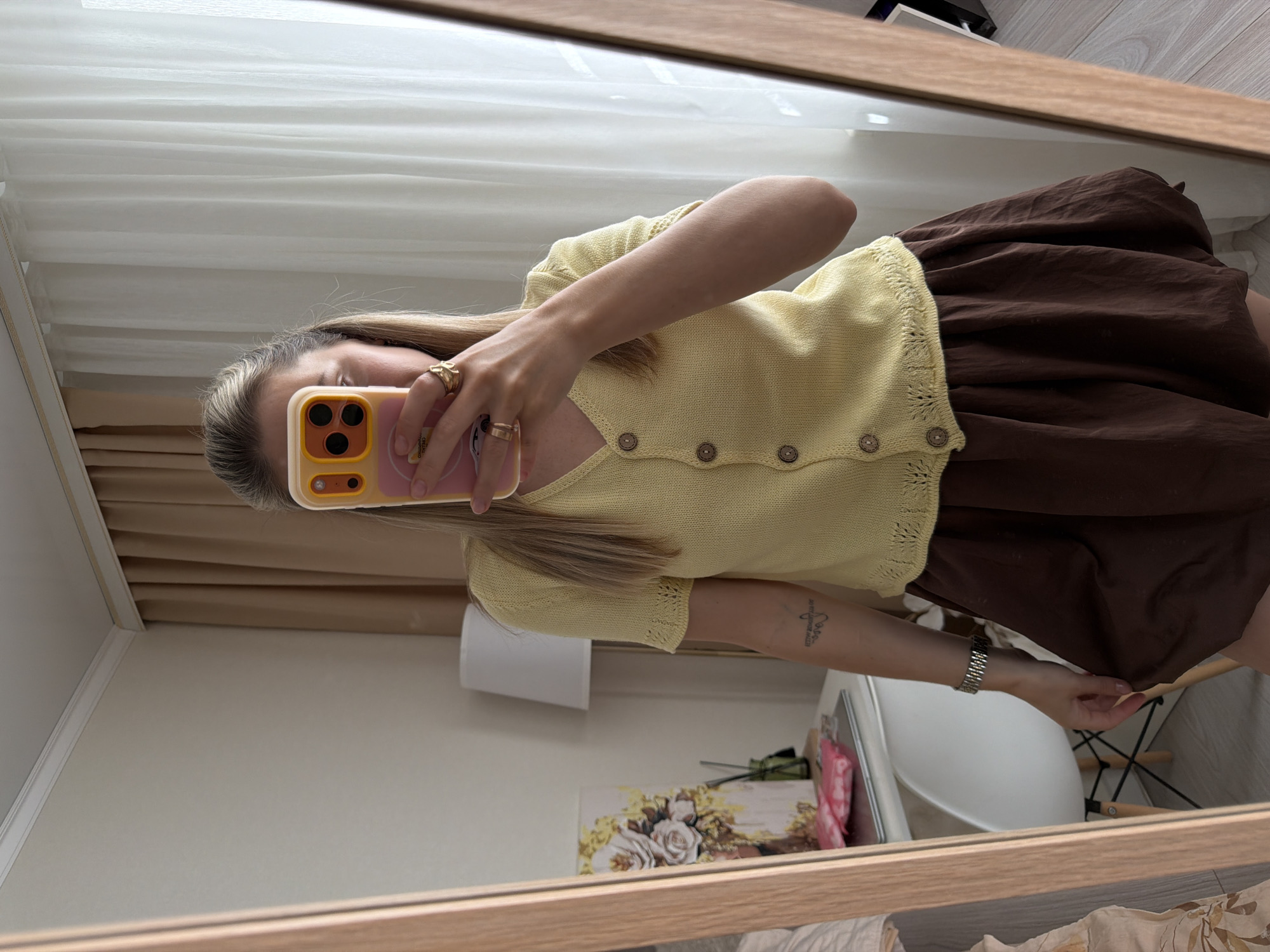 A person is taking a mirror selfie, holding a smartphone in their right hand. They are wearing a light yellow knit cardigan with button closures and a brown skirt. The person has blonde hair and is wearing rings on their fingers. A tattoo is visible on their left arm. In the background, sheer white curtains are visible, along with a glimpse of a lamp and a framed artwork depicting roses. The reflection also shows a white chair and part of a wooden floor. The setting appears to be indoors, possibly a bedroom or dressing area, given the presence of curtains and furniture. The lighting suggests it could be daytime. There is no visible text in the image. The overall mood is casual and relaxed.