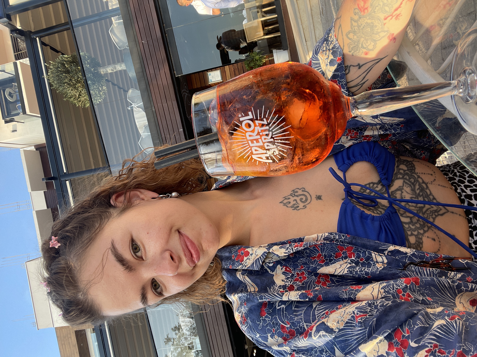 The image shows a young woman smiling and looking at the camera, seated at an outdoor cafe or bar in Budva, Montenegro. She is holding a large glass of Aperol Spritz, which is filled with ice and orange slices, and clearly displays "APEROL SPRITZ" text in white.
The woman has light brown, wavy hair styled with small pink star-shaped hair clips. She wears a blue bikini top under a blue overshirt or kimono patterned with red flowers and white birds (likely cranes or storks). She has several tattoos: a cloud-like design and a small character on her collarbone, and larger, intricate designs on her arms and upper chest/shoulder area. A silver earring with a pearl-like drop is visible on her left ear.
The setting appears to be an elevated outdoor area, possibly a rooftop or balcony, offering views of the sky and nearby buildings. The background features modern architecture with large glass windows and dark frames, reflecting parts of the surroundings, including a person in a white shirt and another in dark clothing. A green, bushy olive tree is visible through a glass partition. The bright, clear blue sky indicates it is daytime with sunny weather. The woman is seated at a glass-top table. The overall scene suggests a relaxed atmosphere, enjoying a drink on a sunny day.