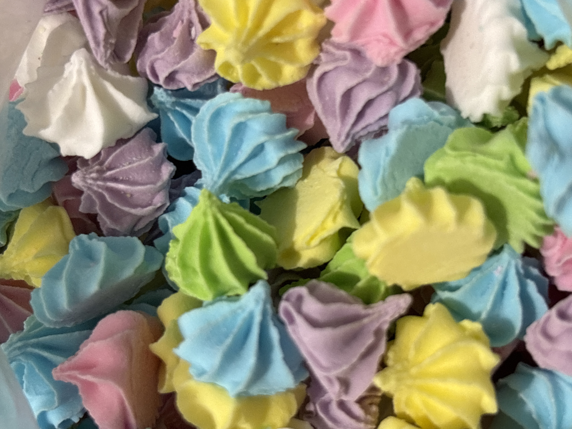 The image shows a close-up, overhead view of a pile of colorful meringue cookies. The cookies are pastel shades of pink, yellow, blue, green, purple, and white. They are piped into a swirled, star-like shape. The lighting is bright and even, and the focus is sharp. There is no indication of the city of Zubov, Russia, in the image. The image is a still photograph of food.