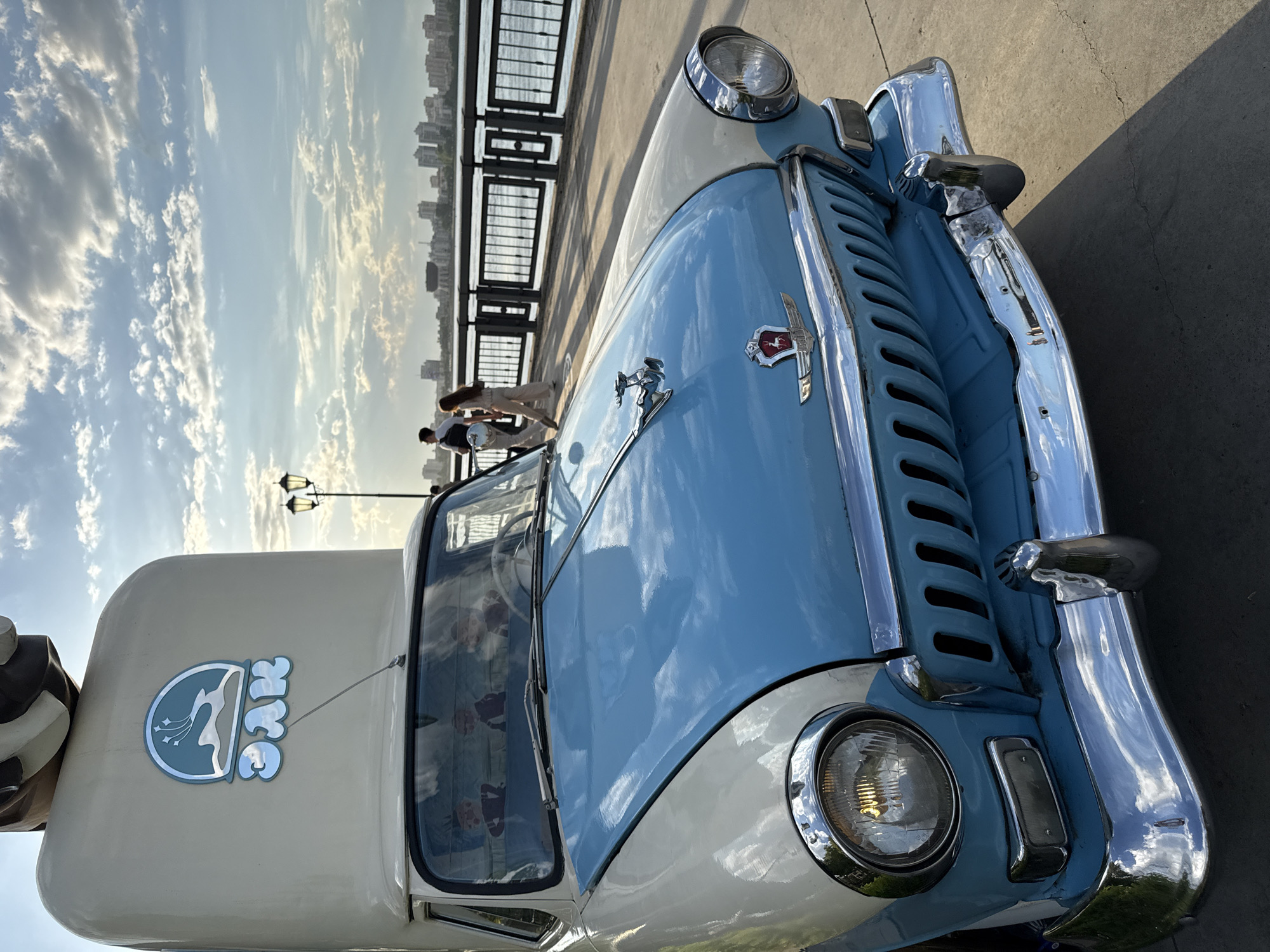 The image shows a vintage car, likely a GAZ-21 Volga, converted into an ice cream truck. The car is painted in two tones: light blue and white. The hood is light blue with chrome details, including the hood ornament. The body is white, and there's a logo on the side featuring a mountain range inside a circle with the text "Эскимо" (Eskimo) above the logo.
In the background, there's a railing with a cityscape visible in the distance. Two people are seen near the railing, possibly customers.
Based on the "Эскимо" logo and architecture, the location is likely Kazan, Russia.