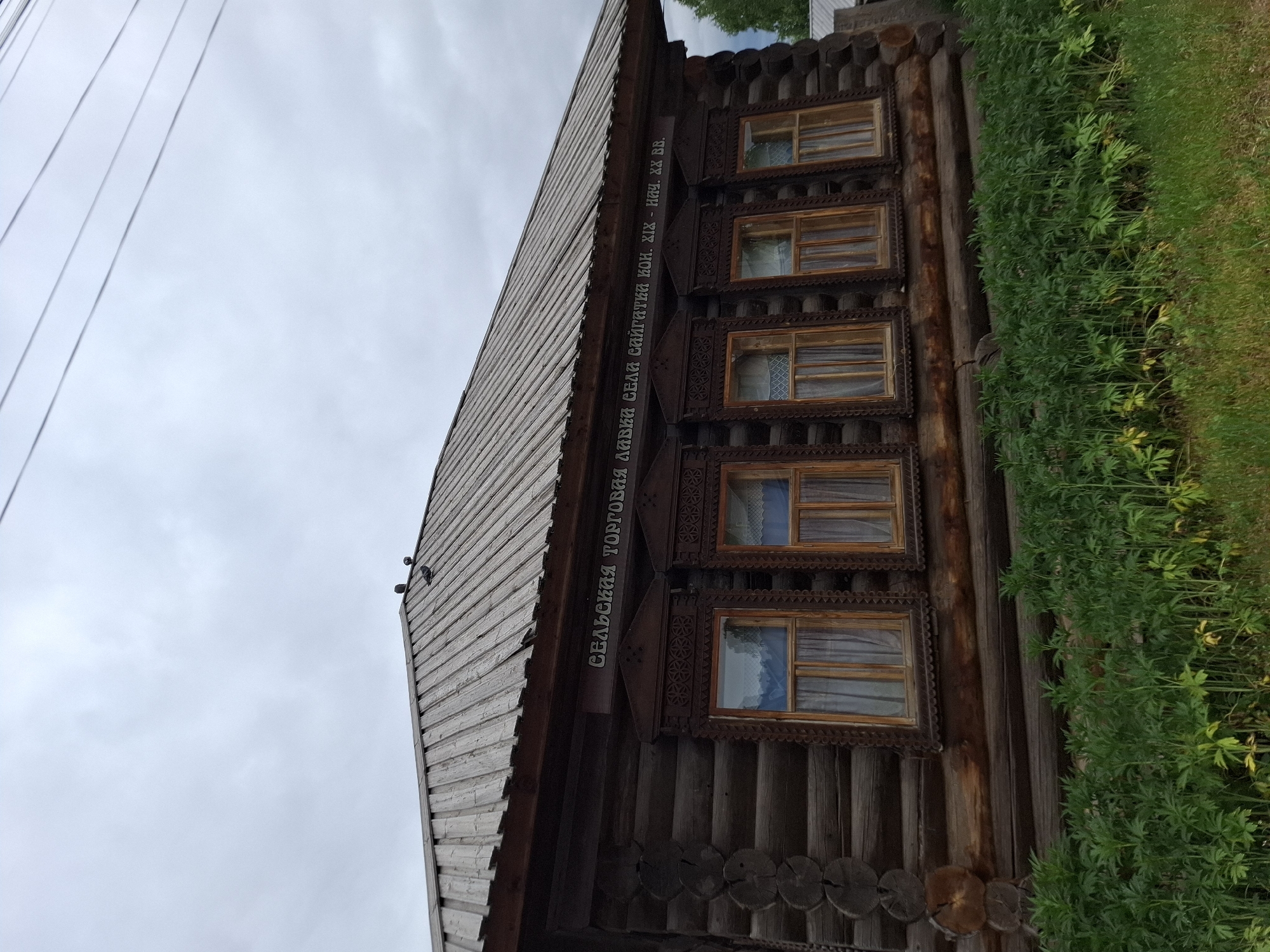 A low-angle shot shows a dark, two-story log cabin structure in what appears to be Chaikovsky, Russia. The structure’s windows are trimmed with decorative wooden latticework, and there are six windows visible in the image. A sign, written in Russian, is located on the facade above the windows. The building's wooden roof is at the left. Lush green vegetation partially obscures the bottom right of the building. Overcast skies fill the background.