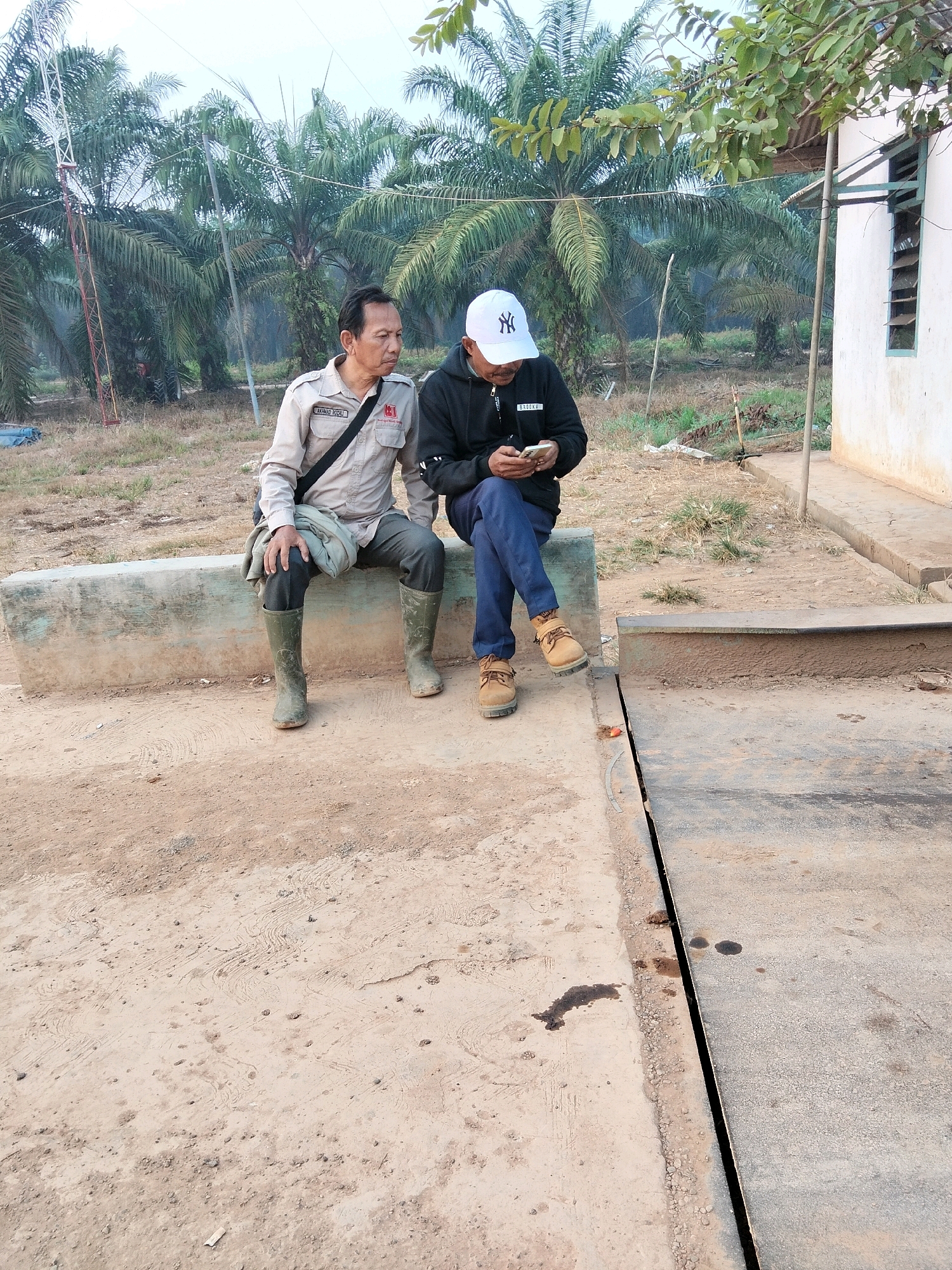 Two men are sitting on a concrete bench in a rural area, likely Teluk Tenggirik, Indonesia, with palm trees in the background. The man on the left is wearing a light-colored collared shirt with an ID badge and dark pants tucked into green rubber boots. He has a light-colored bag slung over his shoulder. The man on the right is wearing a black hooded jacket and blue pants. He is wearing a white baseball cap with a black logo and brown lace-up shoes. He is looking down at a mobile phone in his hands. The ground is dirt and there is a small building to the right of the men.