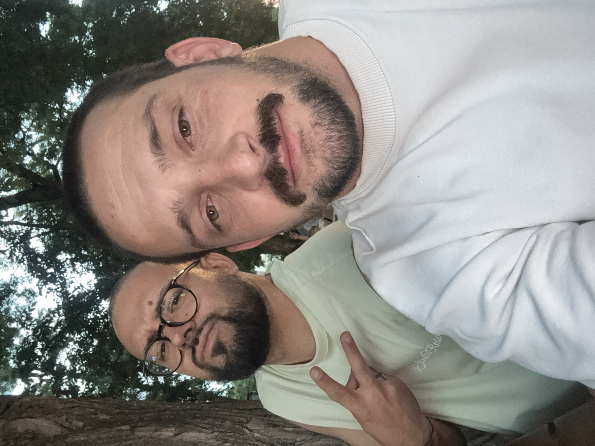 This image, reportedly captured in the city of Plovdiv, Bulgaria, depicts two adult males in a close-up selfie-style photograph taken outdoors.
The subject on the left, positioned behind and slightly to the left of the foreground individual, is a male with dark hair, a trimmed dark beard, and a mustache. He wears dark-rimmed eyeglasses and a light green sleeveless top, on which faint white script text, possibly "Studio Reso," is visible across the chest. His left hand is raised, forming a V-sign (peace sign), and a small black cross tattoo is present on his left ring finger. His facial expression is a slight pout directed towards the camera.
The subject in the foreground, on the right, is a male with dark hair, brown eyes, a prominent dark mustache, and a full dark beard. He is wearing a light-colored, long-sleeved crewneck shirt, which appears to be white or pale blue. His gaze is directed straight into the camera, indicating he is likely holding the camera device.
The background consists primarily of dark green tree foliage, with a vertical tree trunk visible on the far left, suggesting an outdoor natural environment such as a park. The soft, ambient lighting indicates conditions typical of dusk or early evening. No specific urban or architectural landmarks of Plovdiv are discernible within the frame. The interaction between the two individuals appears to be a casual pose for a photograph.