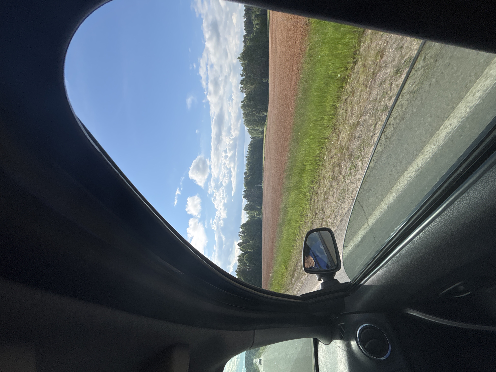 The image is taken from inside a vehicle, looking out the passenger window towards a rural landscape. The foreground shows the interior of the car, including the door panel, a circular vent, and a side mirror reflecting a person wearing a blue top. The window is open, and the view outside includes a paved road with white lines, a grassy verge, a large brown field, and a dense green forest in the distance. The sky is bright blue with scattered white clouds. The scene appears to be from a sunny day, likely in a rural area of Russia given the provided location. The vehicle is in motion, as suggested by the perspective and the blurred road.