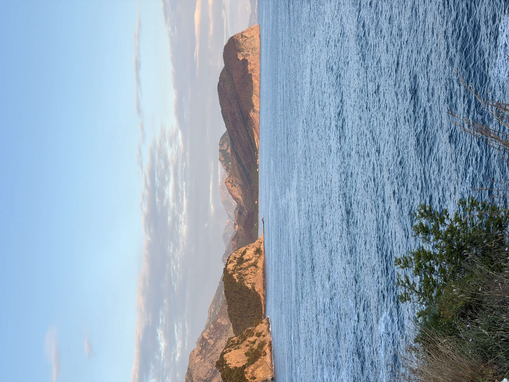 The image presents a view of the Mirište coastline in Montenegro. On the left, a series of reddish-brown, rocky mountains cascade towards the sea. The sky is a pale blue, interspersed with thin, elongated clouds. On the right, the sea, a shimmering expanse of blue, stretches out to the horizon. In the foreground, on the right-hand side, vegetation in shades of green and brown grows on the cliffside. The image is horizontally oriented, showcasing the juxtaposition of land and sea.