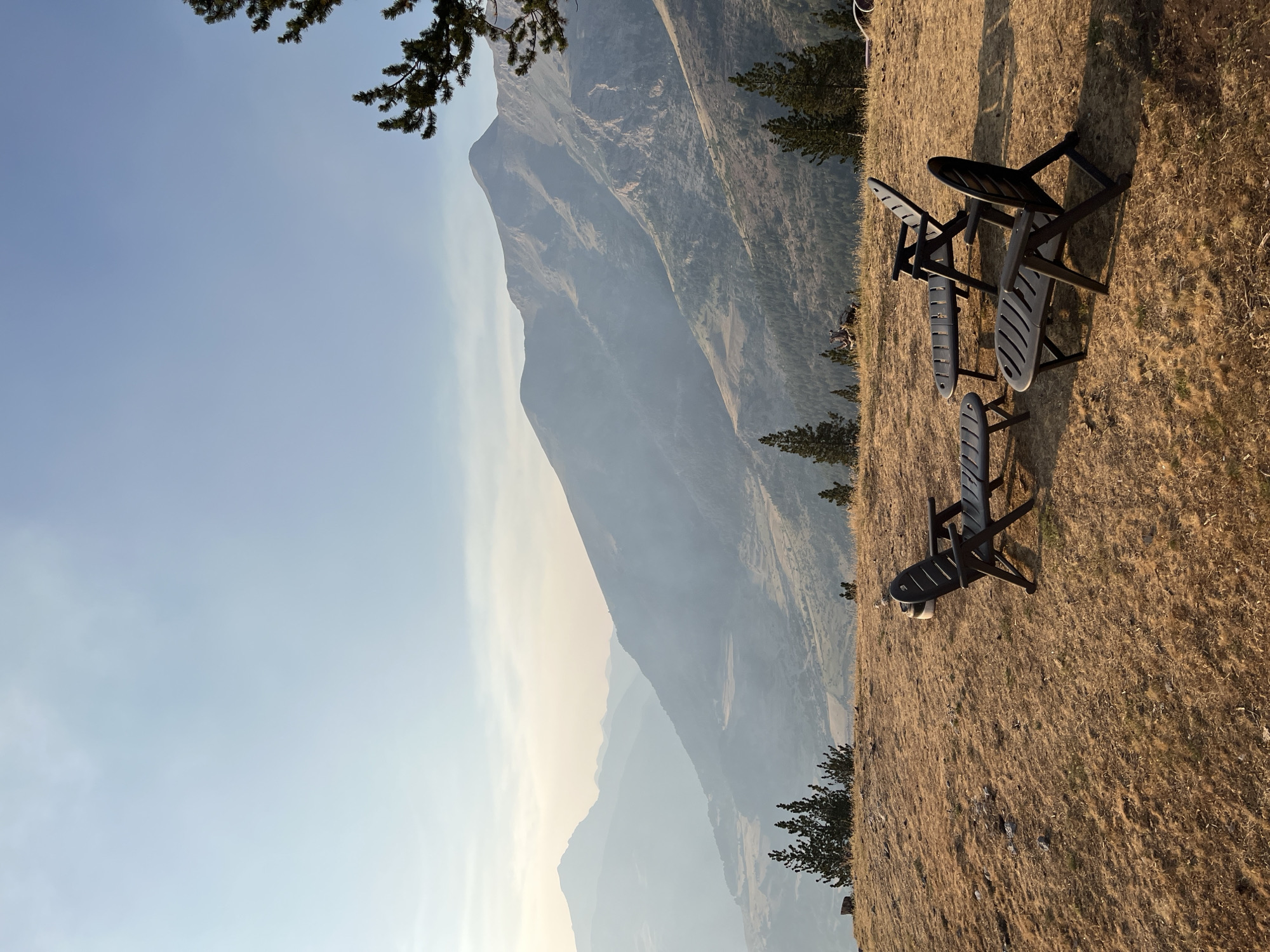The image depicts a scenic overlook in San Juan de Plan, Spain, featuring a mountain landscape. In the foreground, four dark plastic chairs and two small tables are arranged on a dry, grassy hillside. The chairs are positioned to face the expansive view of rolling mountains and valleys, with a hazy atmosphere obscuring the distant peaks. Several pine trees are scattered across the landscape, adding to the natural setting. The sky is a pale blue with faint clouds. There are no people visible in the image. The overall scene suggests a tranquil outdoor seating area designed to appreciate the natural beauty of the mountainous terrain.