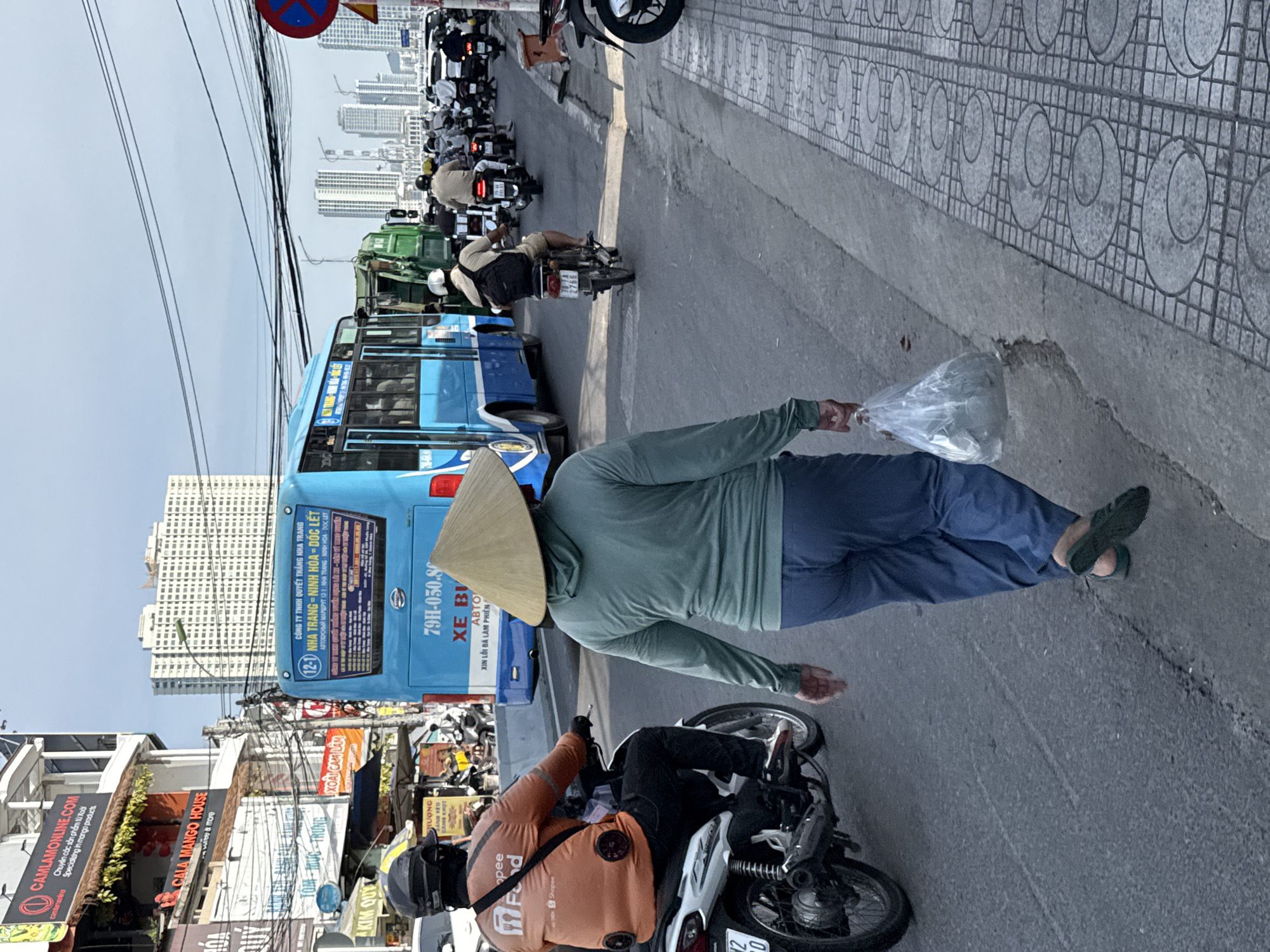 The image depicts a street scene in Bac Nha Trang Ward, Vietnam, characterized by a mix of pedestrian and vehicular traffic under a cloudy sky. In the foreground, a person wearing a conical hat and a green hooded long-sleeved shirt walks away from the camera, carrying a plastic bag filled with items in their right hand. They are dressed in blue pants and dark, flat shoes. To the left, a person on a white motorcycle, wearing an orange long-sleeved shirt with "Shopee Food" branding and a dark helmet, is also moving away from the camera. This motorcycle rider has two circular devices attached to the back of their shirt.
The background is dominated by a bright blue bus with Vietnamese text and route information visible on its rear. Several other motorcycles and a green garbage truck are also present on the road, suggesting a busy urban environment. Buildings line the street, including a multi-story structure with "CAMLAMONLINE.COM" signage. In the distance, tall modern buildings rise against the sky. The road surface is asphalt, and a tiled pedestrian walkway is visible on the right side of the frame. The scene appears to be an ordinary day with ongoing traffic and commercial activity.