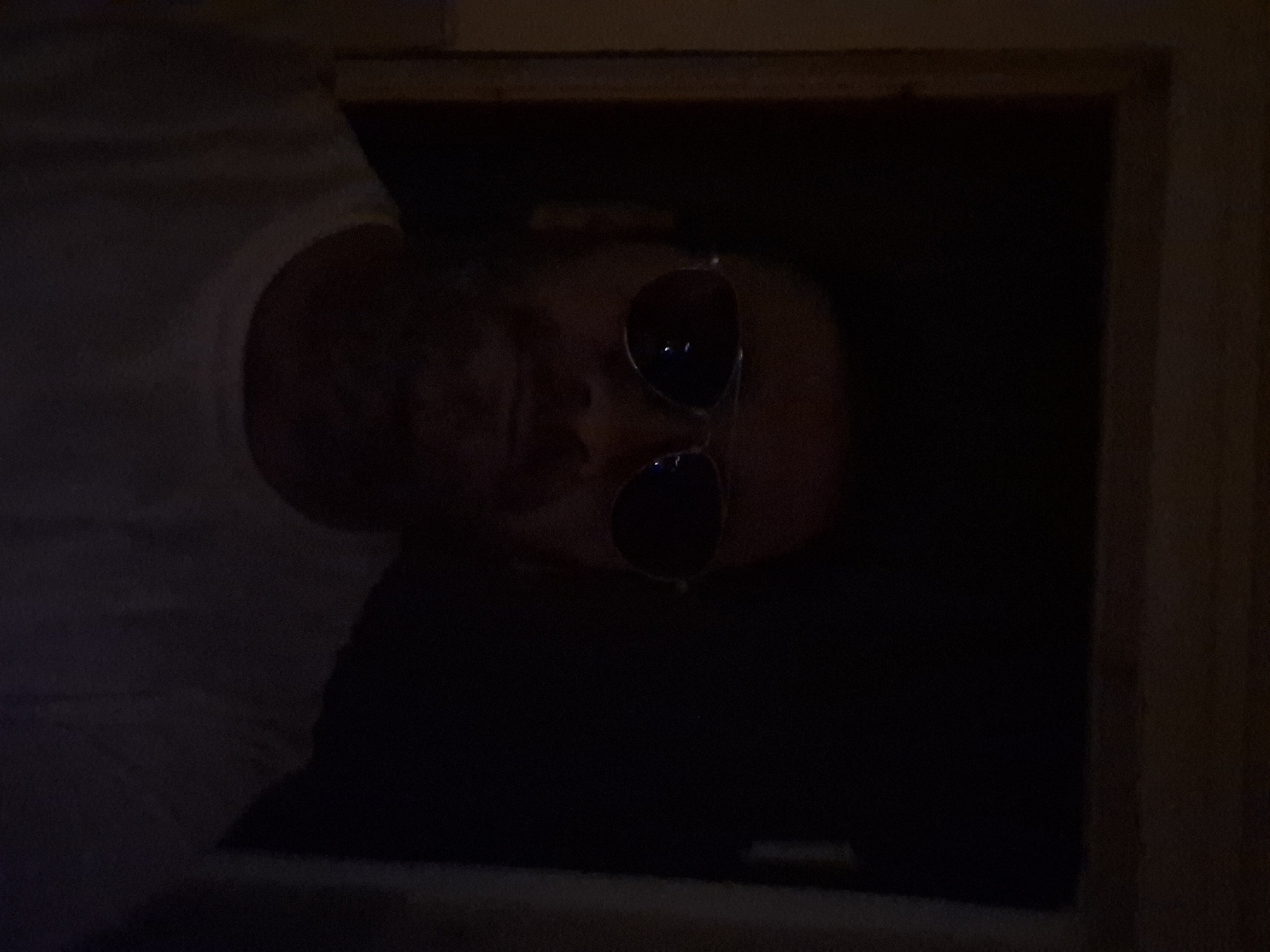 A man is pictured in a dimly lit indoor setting, possibly a doorway. He is wearing sunglasses and a white t-shirt. The background is dark, obscuring most details of the environment. The image appears to be a selfie taken at a low angle. The location is identified as Yerocham, Israel.
