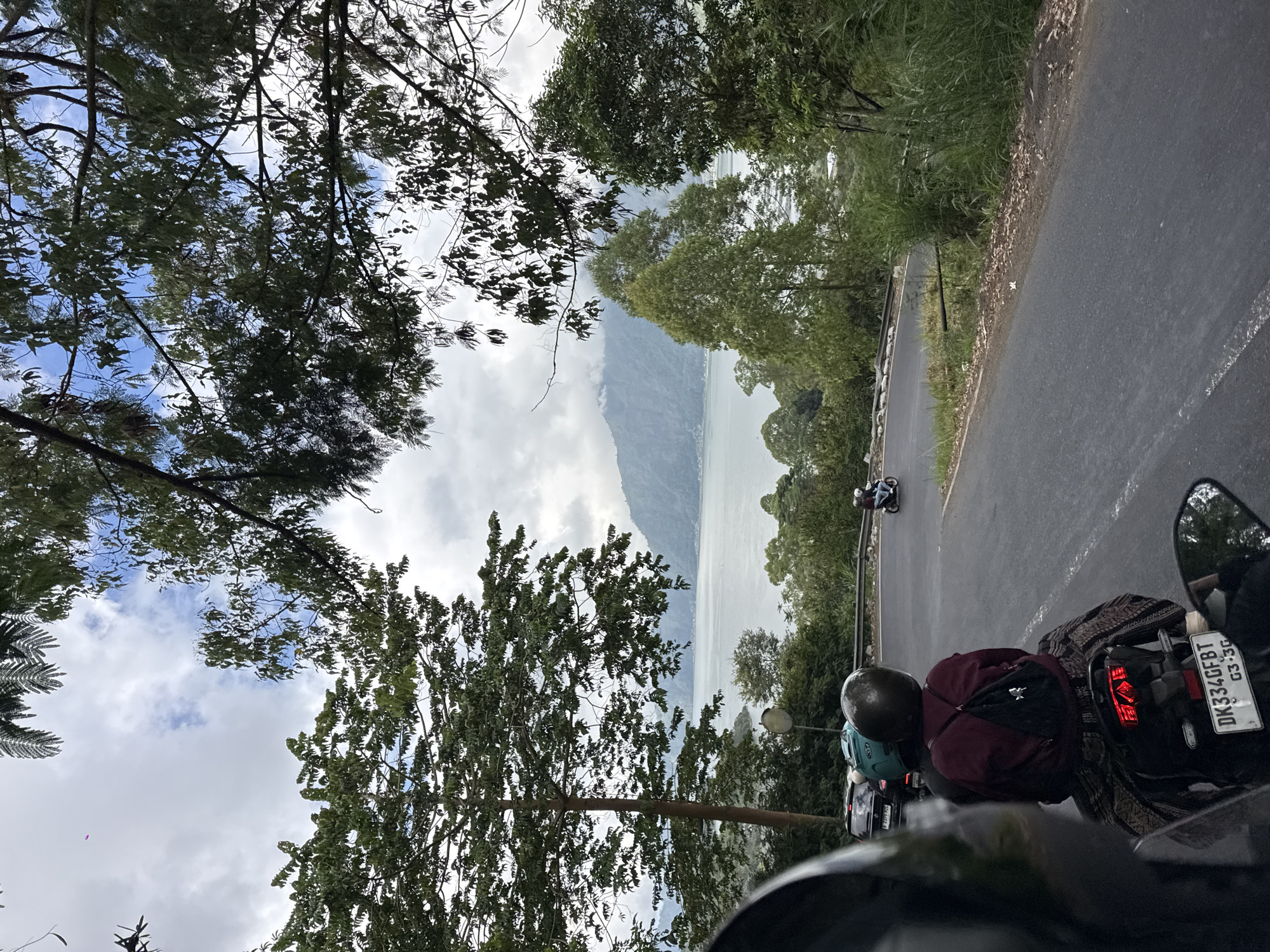 The image depicts a road winding through a verdant landscape with a large body of water and mountains in the distance. The viewpoint is from a motorcycle, with the back of a rider wearing a maroon jacket and black backpack in the foreground. Another motorcycle with a turquoise helmet is visible ahead. The road surface is asphalt, with a white line marking the lane. Vegetation, including trees and tall grasses, lines the sides of the road. In the background, a wide lake is bordered by lush green hills and mist-shrouded mountains, consistent with the scenery of Kintamani, Indonesia, known for its volcanic landscapes and caldera lakes such as Lake Batur. The sky is overcast with patches of blue visible.