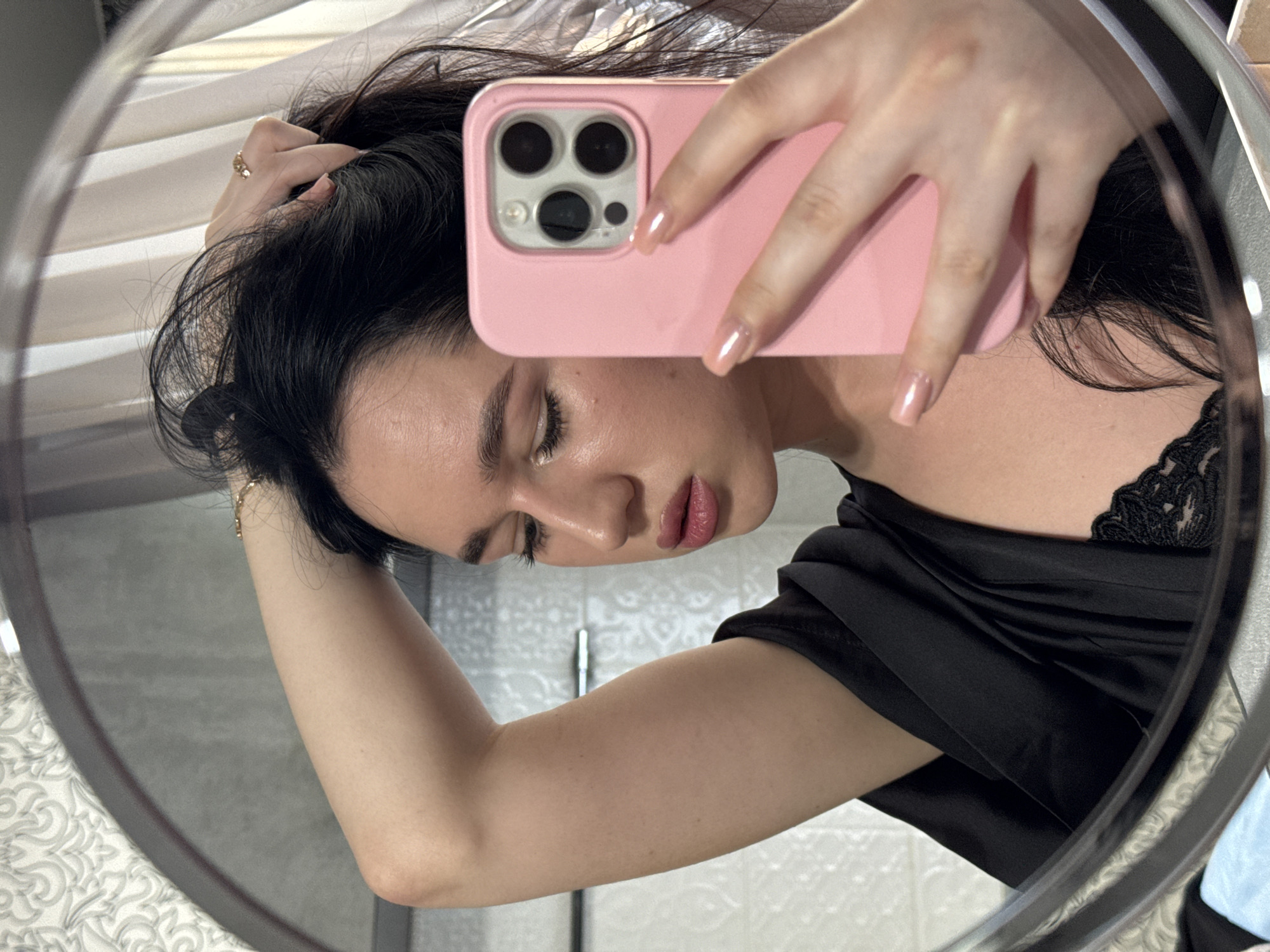 A young person is taking a mirror selfie in a bathroom. The person has dark hair and is wearing a black robe over a black lace bra. Their right arm is raised, and their hand is in their hair. They are holding a pink smartphone in their left hand, which is partially visible in the frame. The background shows a patterned wallpaper, a tiled wall, and a curtain. The image appears to have been taken in a residential setting. There is no information in the image to suggest a location in Novosibirsk, Russia.