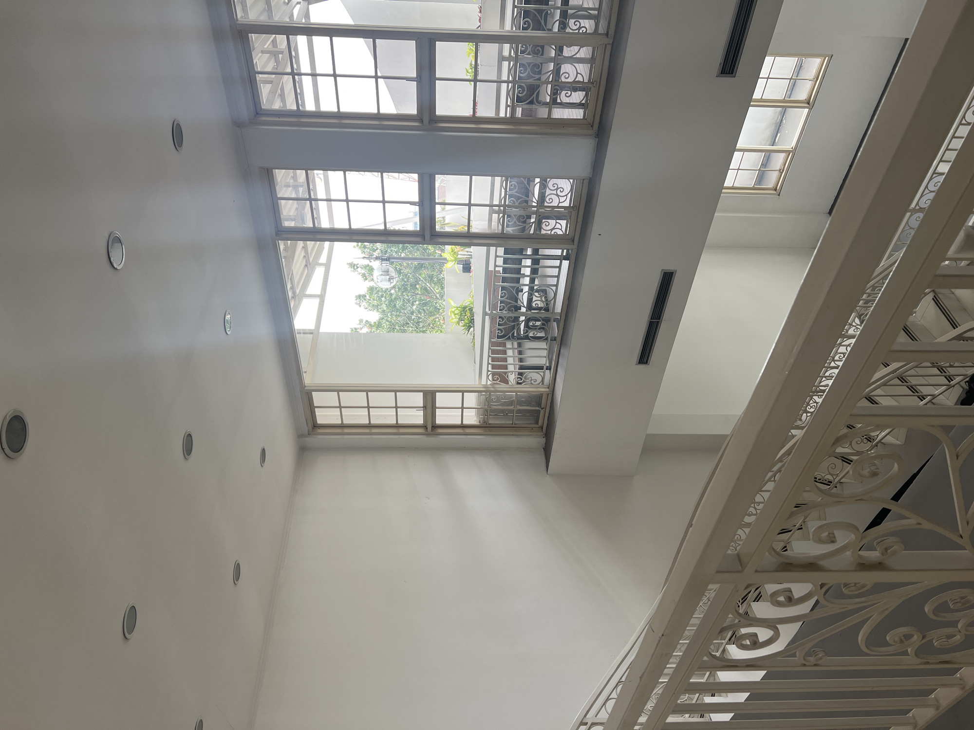 The image depicts an interior view of a building, likely a hotel or a public space, with a prominent staircase and large windows. The staircase, made of white metal with ornate railings, curves downwards, suggesting it leads to a lower level. The walls are painted white, and the ceiling features multiple circular light fixtures.
Large, grid-patterned windows are visible on the upper level, offering a view of the outdoors. Through the windows, greenery and what appears to be a balcony with tables and chairs can be seen. The windows themselves are framed in a light-colored material, possibly aluminum or wood.
The scene is brightly lit, with natural light streaming in through the windows, casting subtle shadows on the walls. There are no people visible in the image. The overall aesthetic is clean, modern, and somewhat minimalist. The presence of air vents near the ceiling suggests an indoor climate-controlled environment. The building's design implies an attempt to maximize natural light and create an open, airy feel.
While the image does not provide specific identifiers for Bandung City, Indonesia, the architectural style and the type of greenery visible outside could be consistent with urban environments in Indonesia. Without further context or specific landmarks, it is impossible to pinpoint an exact location within Bandung City.