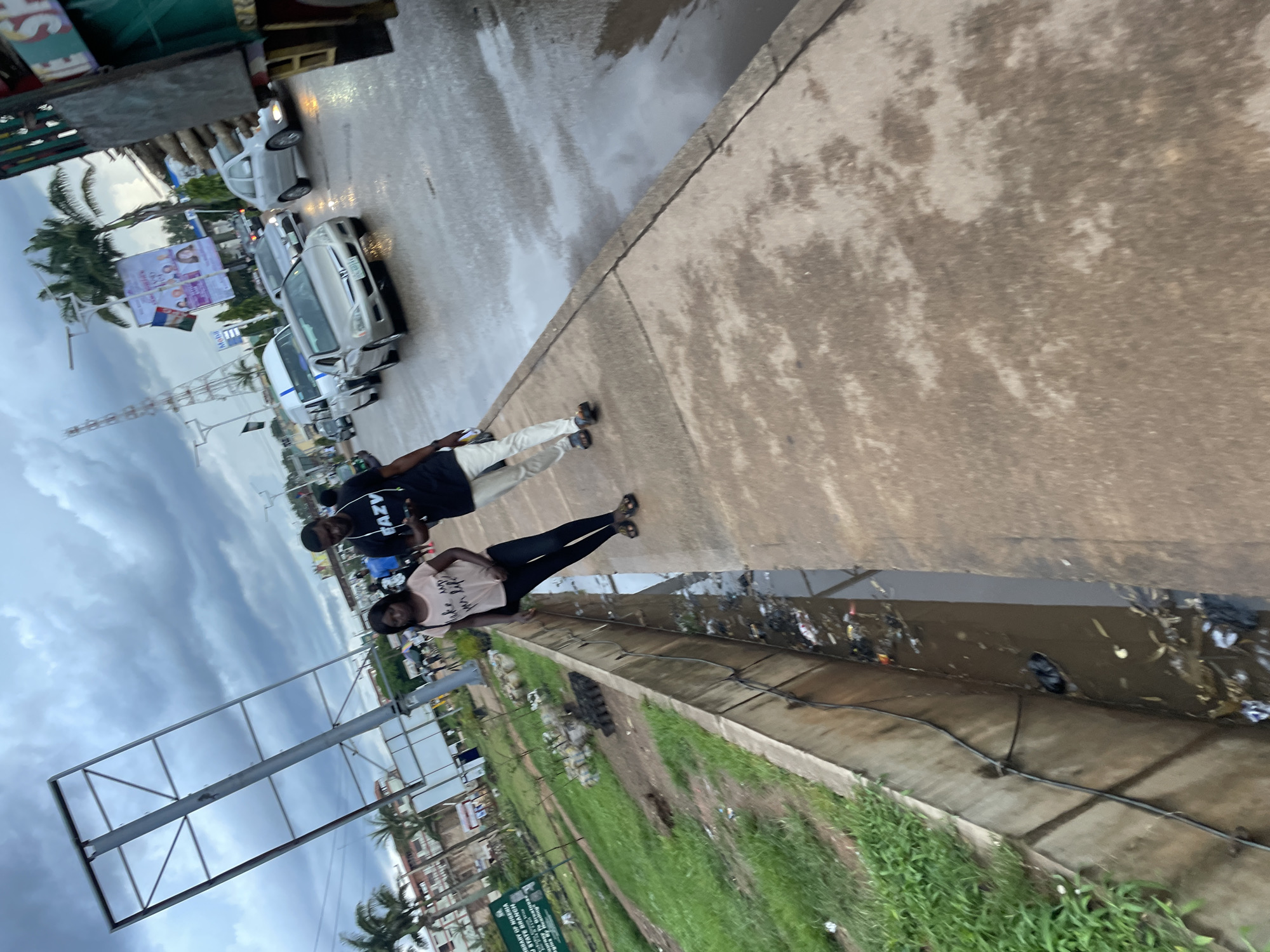 A couple is walking on a sidewalk next to a dirty drainage ditch in Jos, Nigeria. The man is wearing a black t-shirt with "RAZY" written on it, and white pants. The woman is wearing a pink t-shirt and black leggings. Cars are driving on a wet road beside the sidewalk, and there are palm trees and buildings in the background. The sky is cloudy.