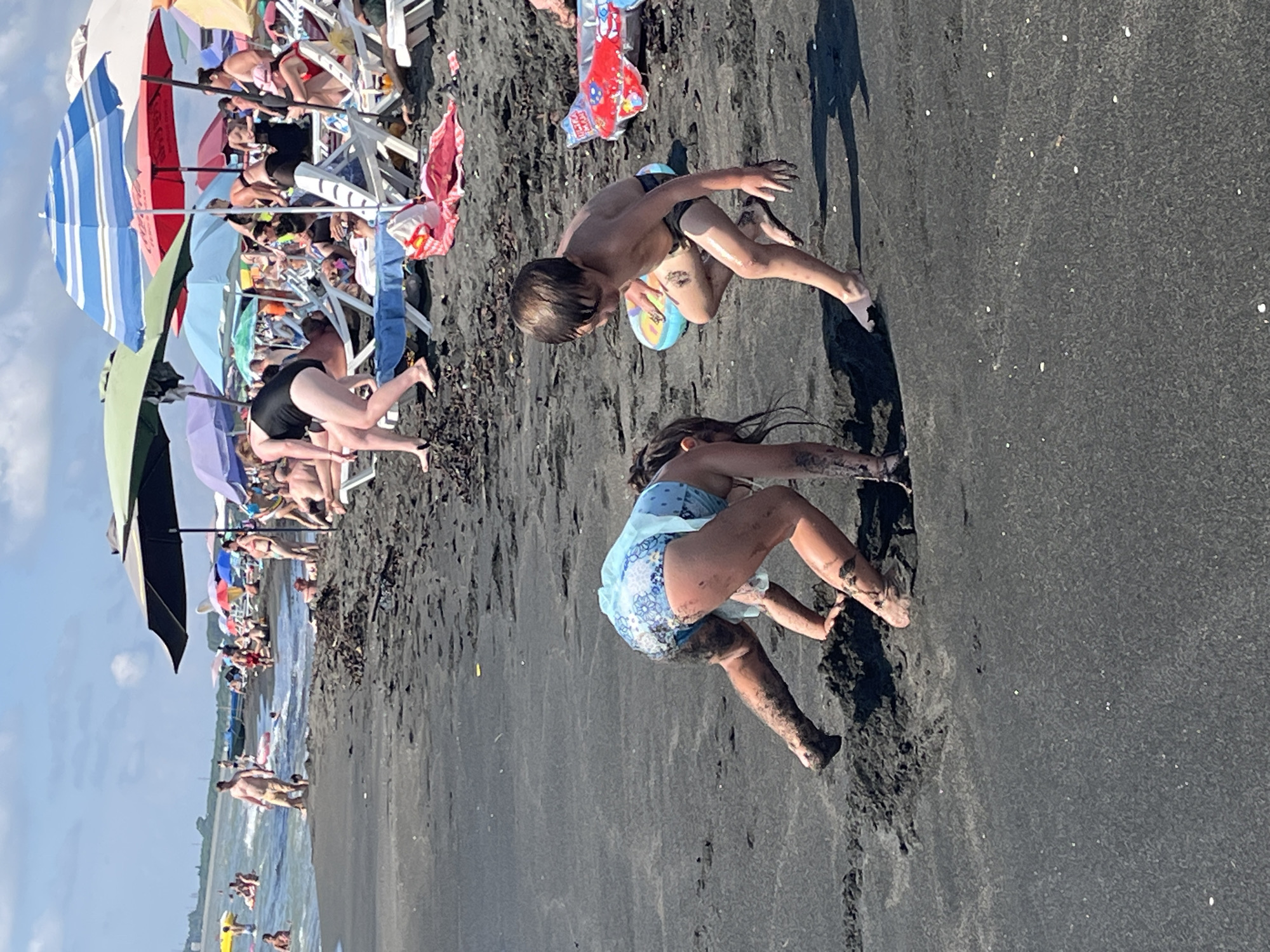 In Ureki, Georgia, a beach scene unfolds on a sunny day with black sand. Two children are the primary subjects in the foreground, engaged in play. A girl, wearing a blue swimsuit with a floral pattern, is on her hands and knees, covered in wet sand, with her back to the camera. Beside her, a boy, wearing dark swim trunks, is crouched down, looking towards the girl, with wet sand on his legs and torso. He holds a colorful circular toy.
In the mid-ground, numerous beachgoers are scattered across the dark sand, many relaxing on lounge chairs under various brightly colored umbrellas, including green, blue, red, and striped designs. Some people are in the shallow water, while others are walking along the shoreline. The water appears calm and blue. The overall environment suggests a populated and active beach setting.