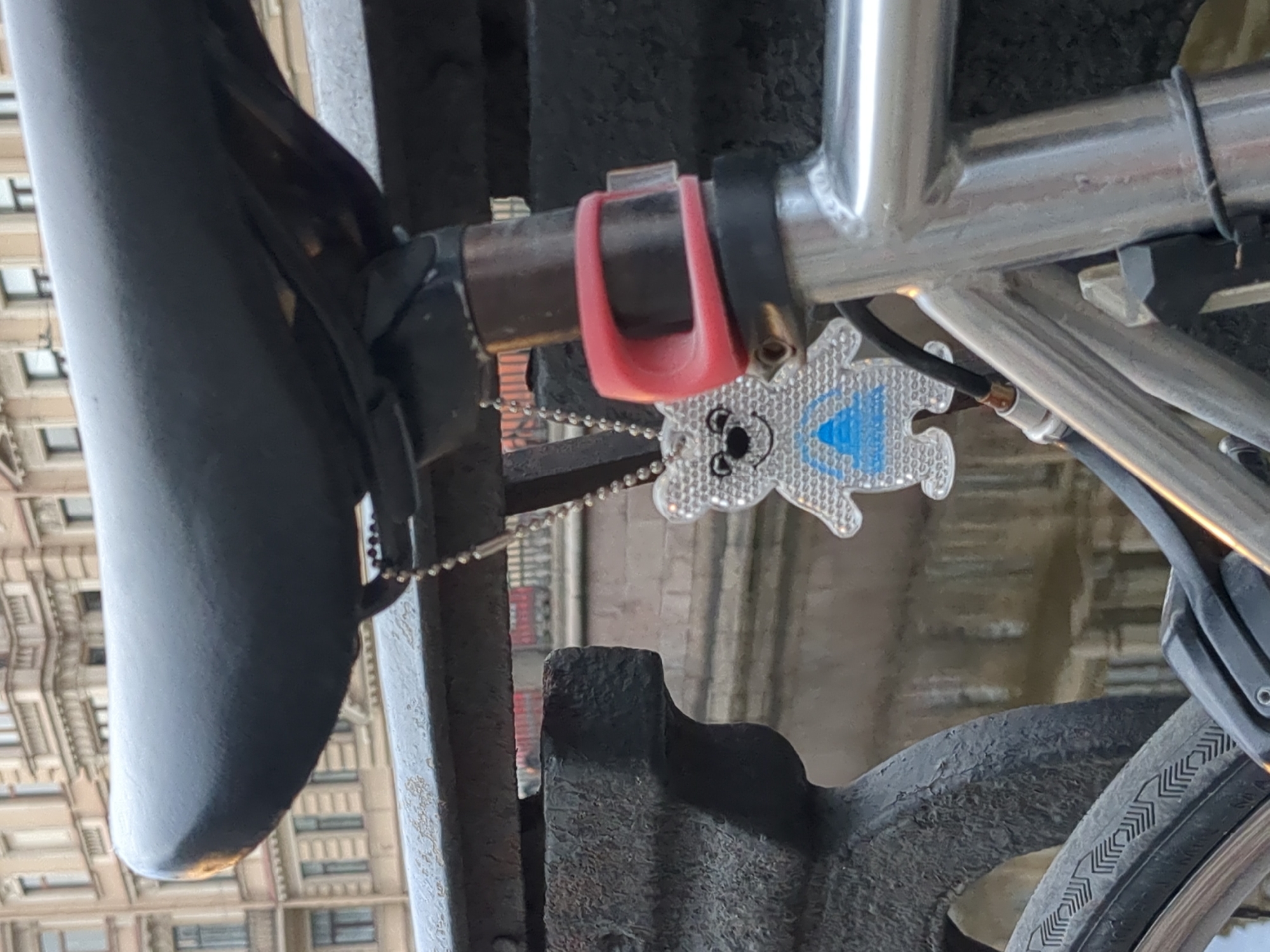 A close-up photograph depicts a bicycle parked outdoors in Saint Petersburg, Russia. The image focuses on the bike seat, frame, and rear tire, with a building visible in the blurred background. A reflective bear-shaped charm hangs from the seat post. A red silicone ring is attached to the frame.