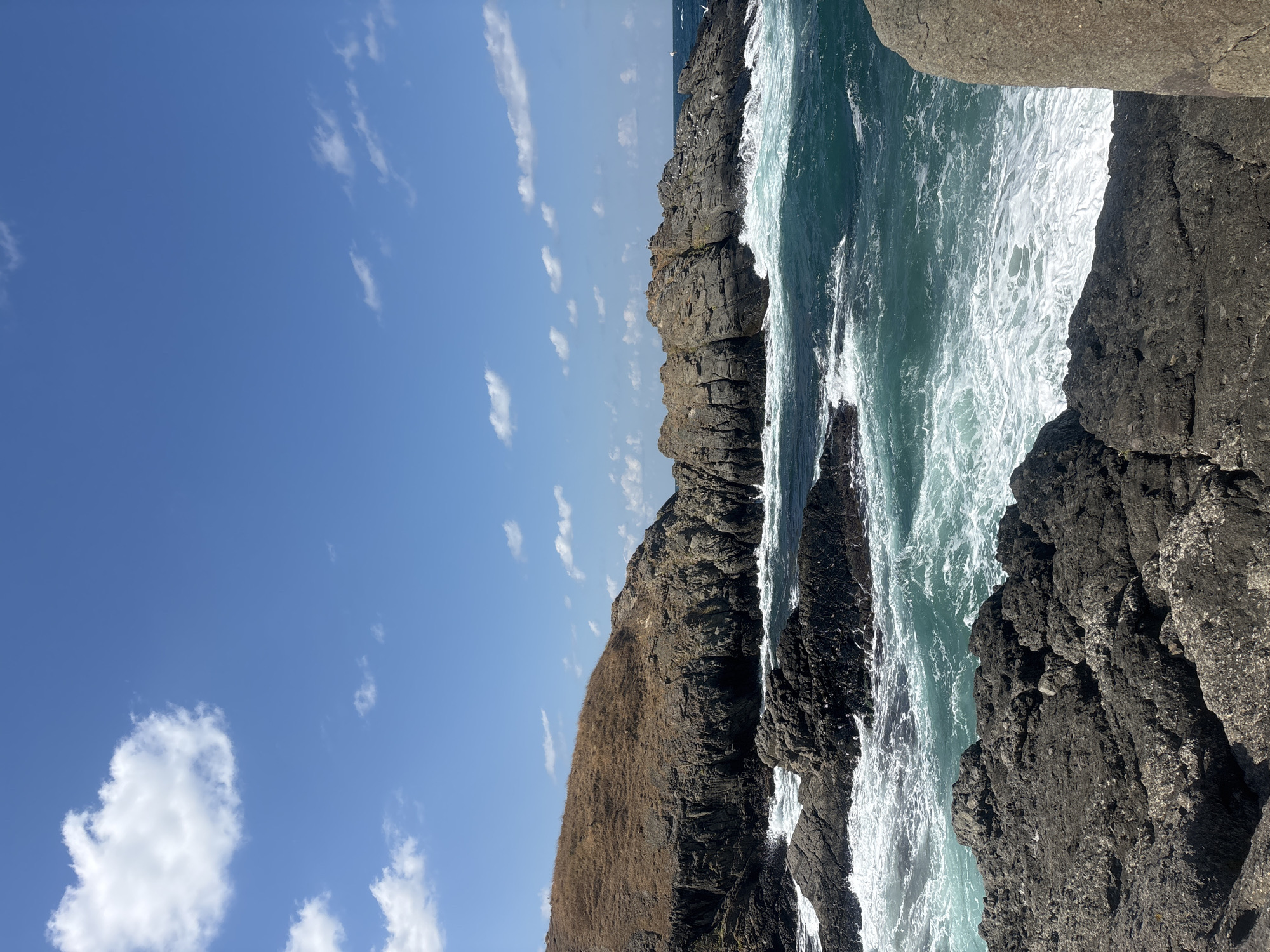 This is a photograph of the rocky coastline near the town of Sinemorets, Bulgaria. The image captures a dynamic scene with rough, dark grey rocks in the foreground and middle ground, jutting out into the turquoise sea. Waves crash against the rocks, creating white foam and spray.
In the background, a large, undulating hill covered in dry, golden-brown grass rises above a dark, sheer cliff face. The cliff is rugged and appears to be composed of many smaller rock formations. The sea is a vibrant teal color, with whitecaps indicating choppy conditions. The sky above is a clear, bright blue, dotted with a few fluffy white clouds.
There are no people or animals clearly visible in the image, although a few small white specks that could be birds are present near the rocks in the middle distance. The overall impression is one of natural, rugged beauty and the power of the sea. The image was likely taken during the daytime, under sunny conditions.
