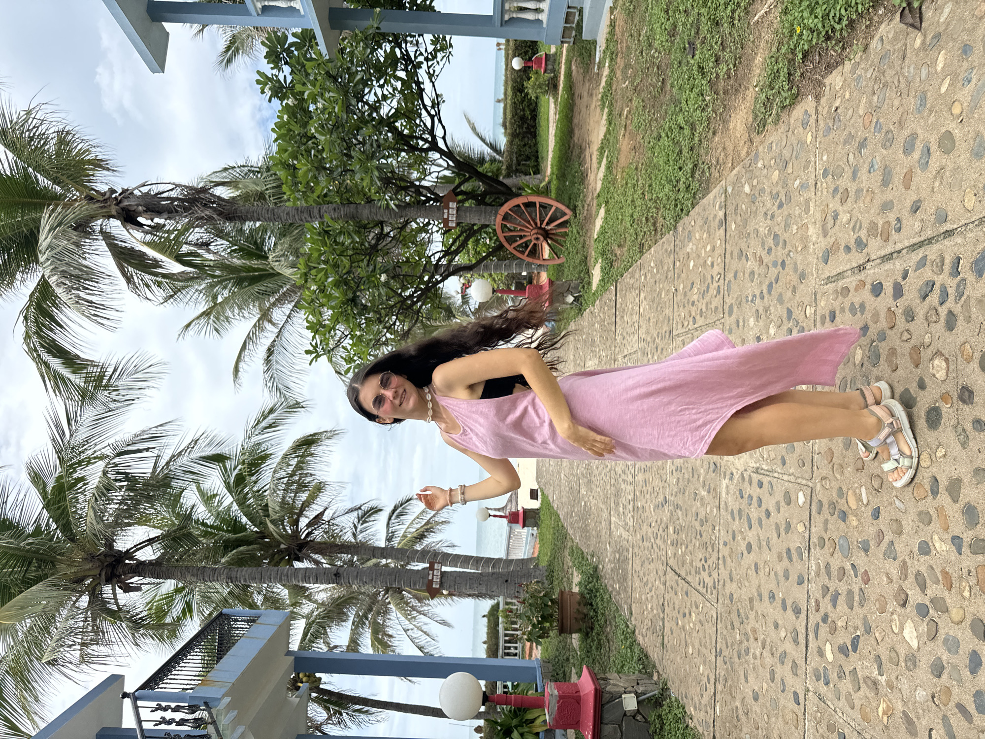 An outdoor, rotated (sideways) full-shot photograph features a young woman smiling at the camera on a paved path in a tropical setting. The woman has long, dark hair, worn loose and flowing. She wears a light pink, sleeveless maxi dress over a dark top, round pink-tinted sunglasses, a shell necklace, and light-colored sandals with pastel straps. Her arms are relaxed, with one hand slightly raised.
The setting appears to be a resort or tropical garden in Mũi Né, Vietnam, as indicated by a small wooden sign attached to a palm tree that clearly reads "MUI NE". Tall palm trees dominate the scene, with their fronds reaching towards a partly cloudy sky. The path she stands on is made of concrete embedded with numerous small, multi-colored pebbles.
To the left, parts of a light blue and white building with decorative railings are visible. To the right, a grassy area with some shrubs runs alongside the path. Further in the background, beyond more greenery and decorative elements like a large wooden wagon wheel leaning against a tree and white spherical lights on red pedestals, the ocean can be seen under the overcast sky. The overall atmosphere is relaxed and vacation-like, with the woman appearing happy and at ease. The time of day is daytime, with soft, diffused lighting.