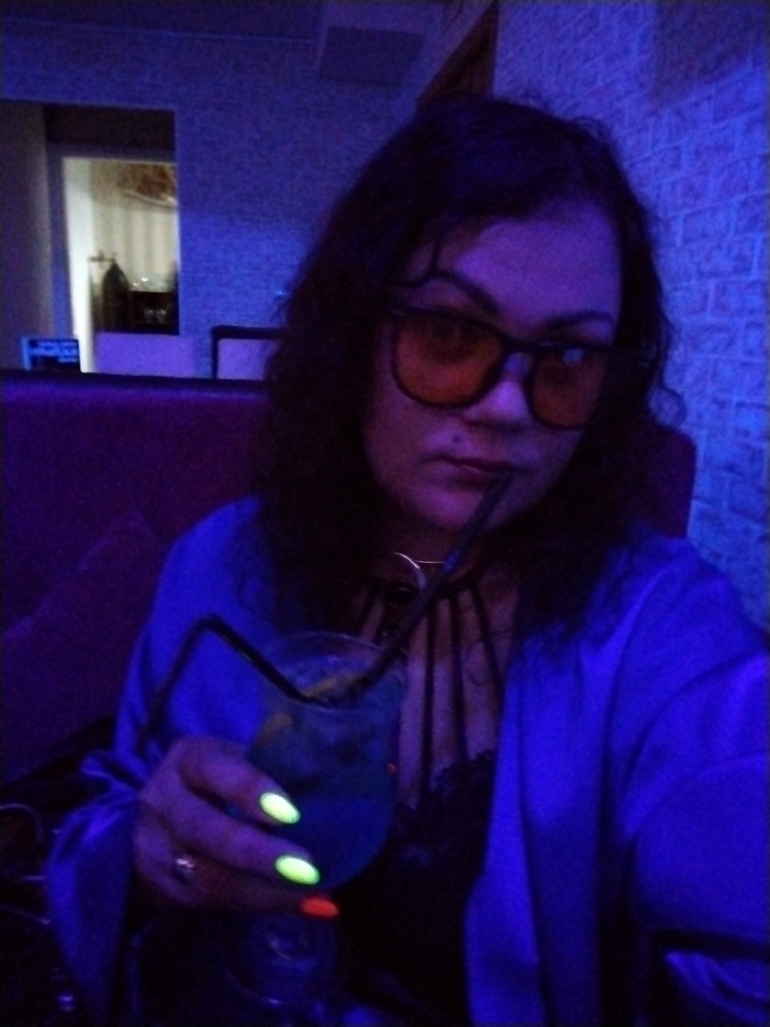 An adult woman is depicted in a selfie or close-up shot, seated in a dimly lit indoor establishment, likely a bar or lounge in Chaikovsky, Russia. The scene is predominantly bathed in purple and blue ambient lighting, giving everything a cool, vibrant hue. The woman has dark, wavy hair and is wearing dark-rimmed glasses with distinctive amber-tinted lenses, which contrast with the blue lighting.
She is dressed in a blue top or jacket, possibly made of a satin-like material, worn over a dark, potentially lace, garment. Around her neck, she has a dark choker-style necklace with multiple decorative straps. The woman holds a clear glass containing a blue-hued drink, likely a cocktail, with a dark straw positioned at her mouth, suggesting she is about to take a sip or has just done so. Her left hand is visible, featuring a ring on her ring finger. Her fingernails are painted in striking colors: neon green/yellow on her thumb, index, and middle fingers, and a contrasting red or orange on her ring finger.
In the background, a textured wall resembling brickwork is visible on the right, appearing blue under the lights. To her left, there is a dark, possibly upholstered, partition or seating. Further back, an opening reveals a glimpse into a brighter adjacent room with striped wallpaper and some indistinct objects, including a dark bottle. A small blue sign with unreadable light-colored text is partially visible on the left side of the background. The overall atmosphere suggests an evening or nighttime social event. Her expression is neutral as she looks towards the camera.