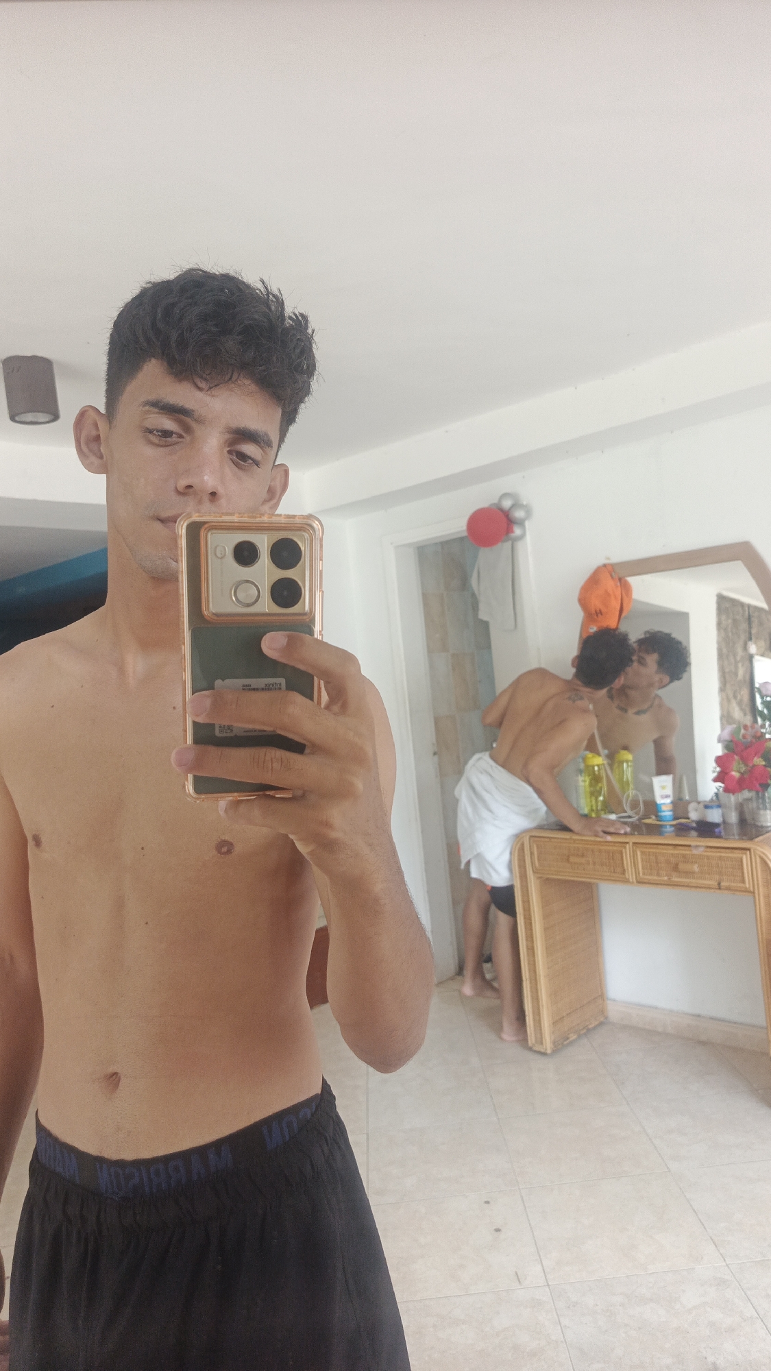 This indoor full-shot image captures a young man taking a mirror selfie, revealing a secondary scene in the reflection.
In the foreground, a light-skinned young man with dark, curly hair and a neutral expression holds a smartphone in his right hand to capture the image. He is shirtless and wears black shorts, with the word "MARRIAM" visible in reverse on the waistband. The smartphone has a distinct gold-colored protective case and a large camera module with four visible lenses/sensors.
The background, primarily seen through a large rectangular mirror with a wooden frame, shows another young man. This person, also shirtless and wearing white shorts or a towel wrapped around his waist, is leaning over a light brown wicker-style dresser or table. He appears to be interacting with his own reflection, leaning into it as if kissing it.
On the dresser, various items are visible, including two green transparent water bottles, a white tube (possibly lotion or sunscreen), and a small vase holding artificial red and pink flowers. An orange cap hangs on the wall beside the mirror. Behind the dresser, a doorway leads to another room, and a cluster of red and silver balloons or decorations is attached to the wall. The room has light-colored tiled flooring and white walls, with a dark gray ceiling light fixture visible in the upper left.
The scene suggests a casual setting, possibly a bedroom or common area in a home or vacation rental in a warm climate, consistent with Playa El Agua, Venezuela. The activity is a selfie being taken, capturing a playful or self-reflective moment in the background. The time of day appears to be daytime due to ample ambient light.